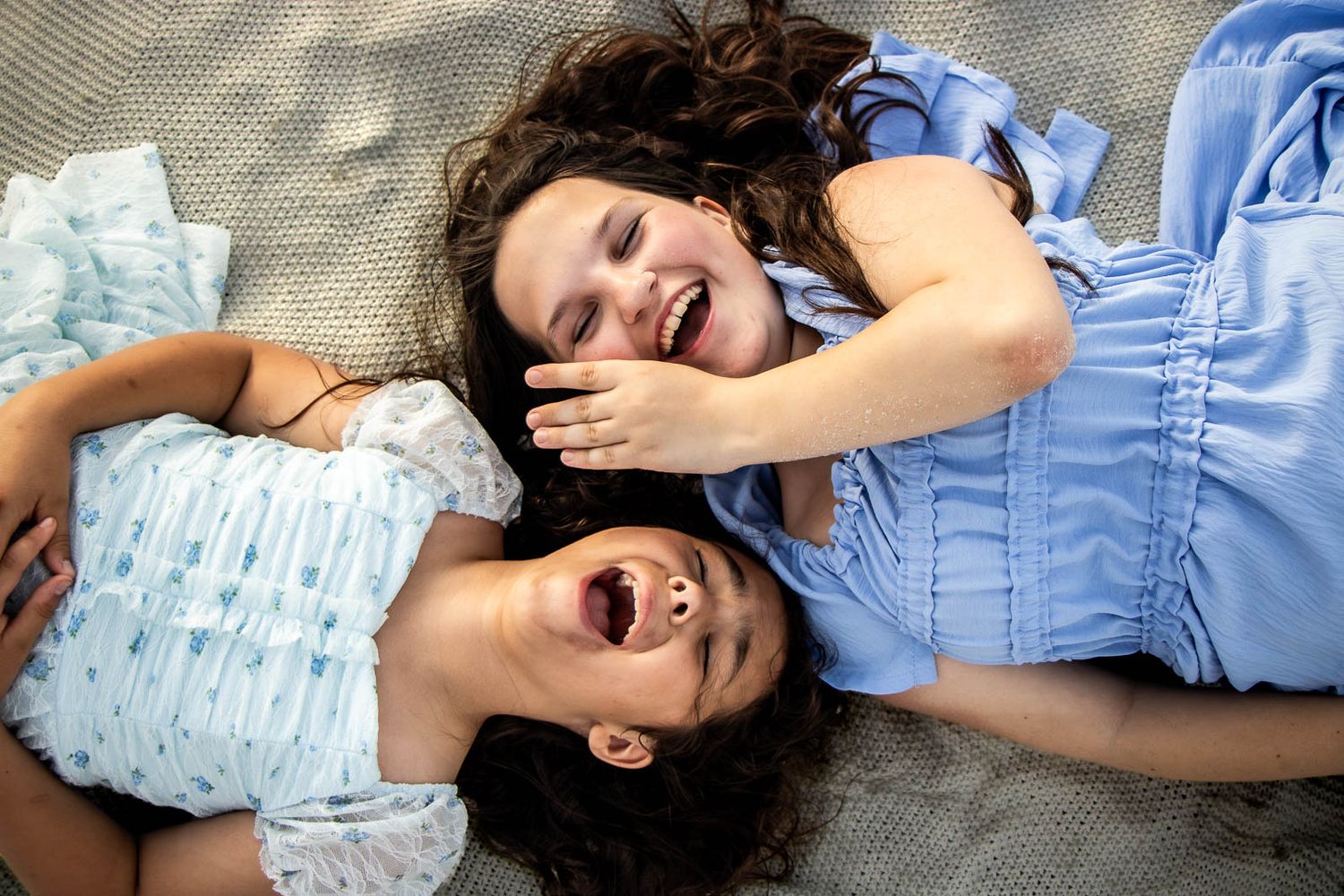 Two young girls laughing and lying on a blanket, wearing light blue dresses, sharing a joyful moment together.