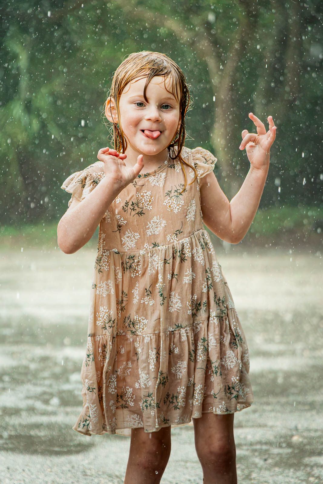 A joyful little girl in a floral dress playing in the rain, smiling with her tongue out and hands raised.