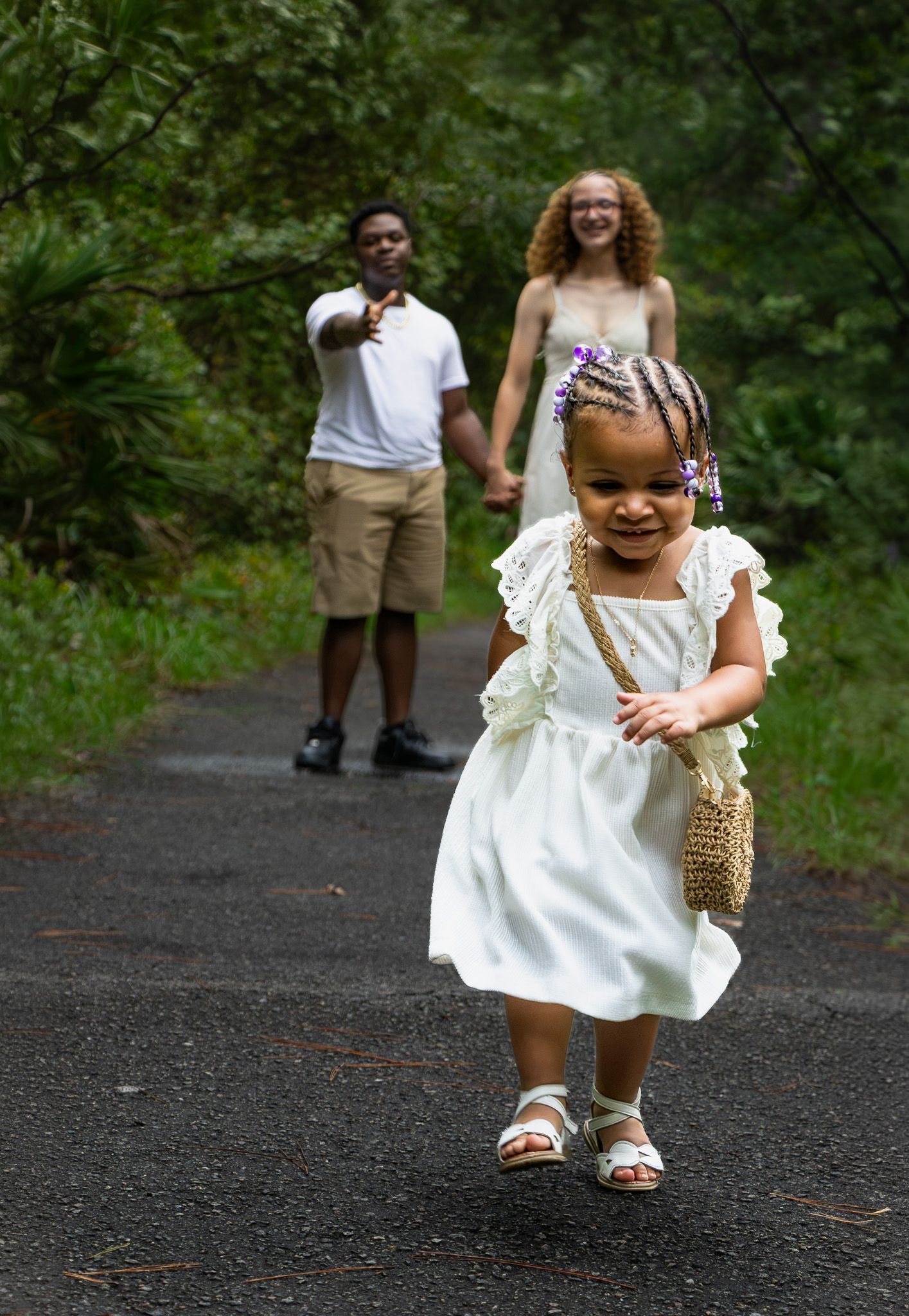 A young girl in a white dress runs down a tree-lined path as her parents hold hands and smile behind her.