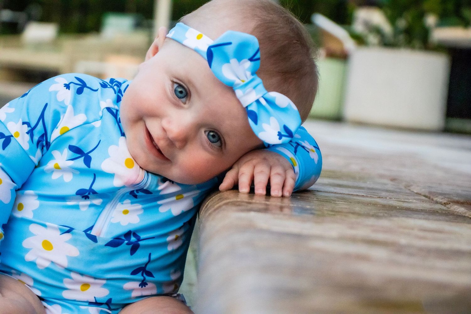 A baby wearing a blue flower swim suit laying her head on a pool and smiling with a bow in her hair.