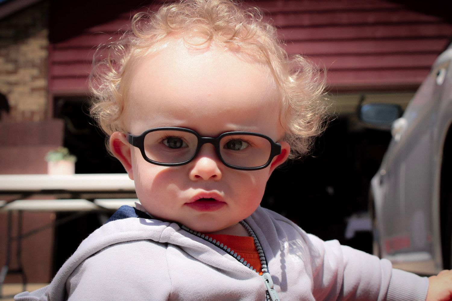 A small child wearing glasses and holding onto a car while standing up with curly hair.