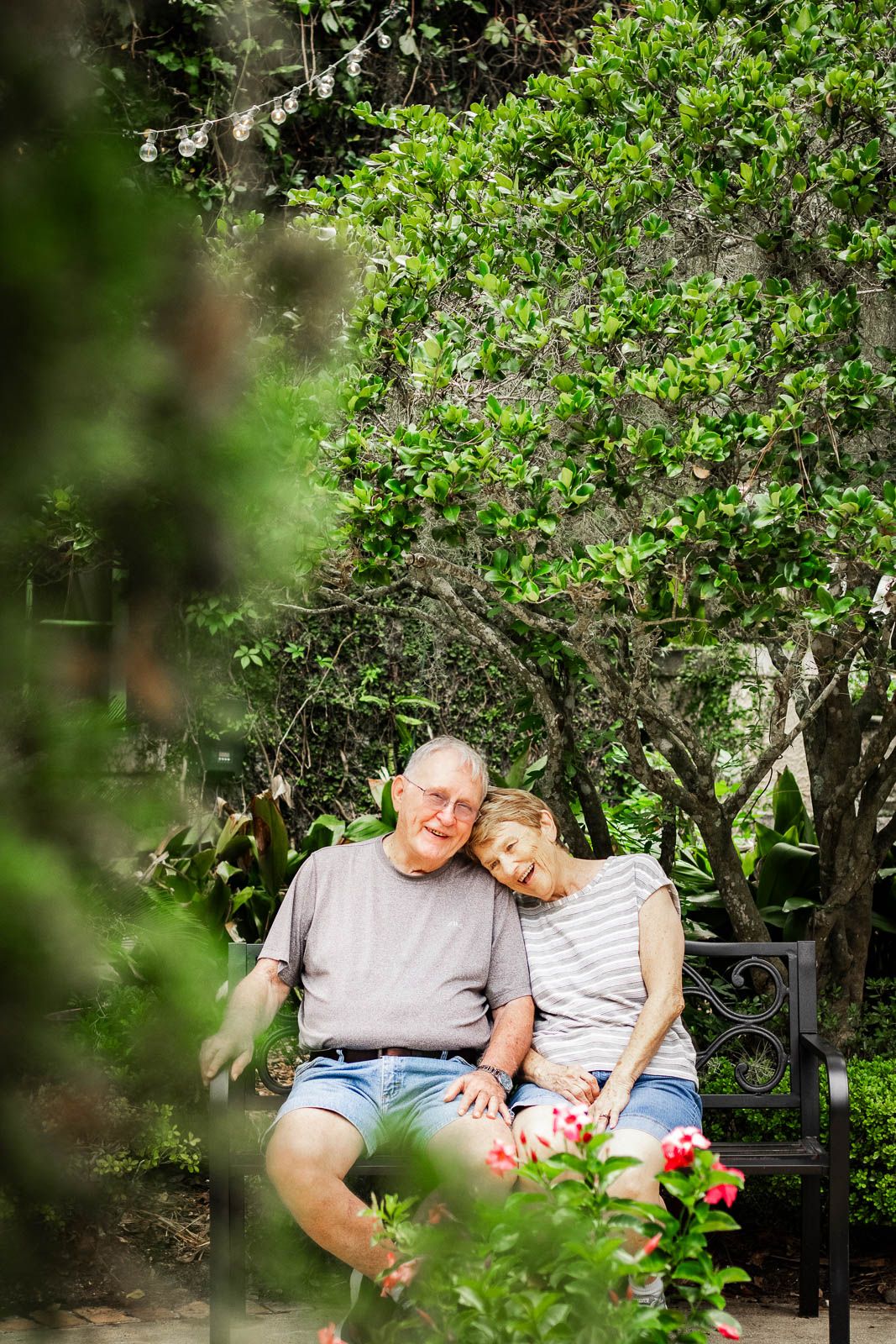 Elderly couple sitting close together on a bench surrounded by lush green trees and flowering plants for their professional photography.