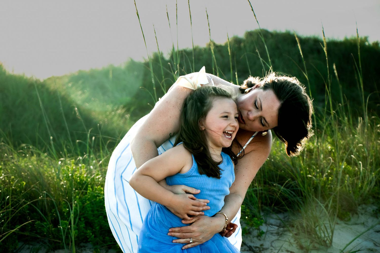 A woman and young girl laughing and embracing joyfully on a sandy beach with tall green grass behind them.