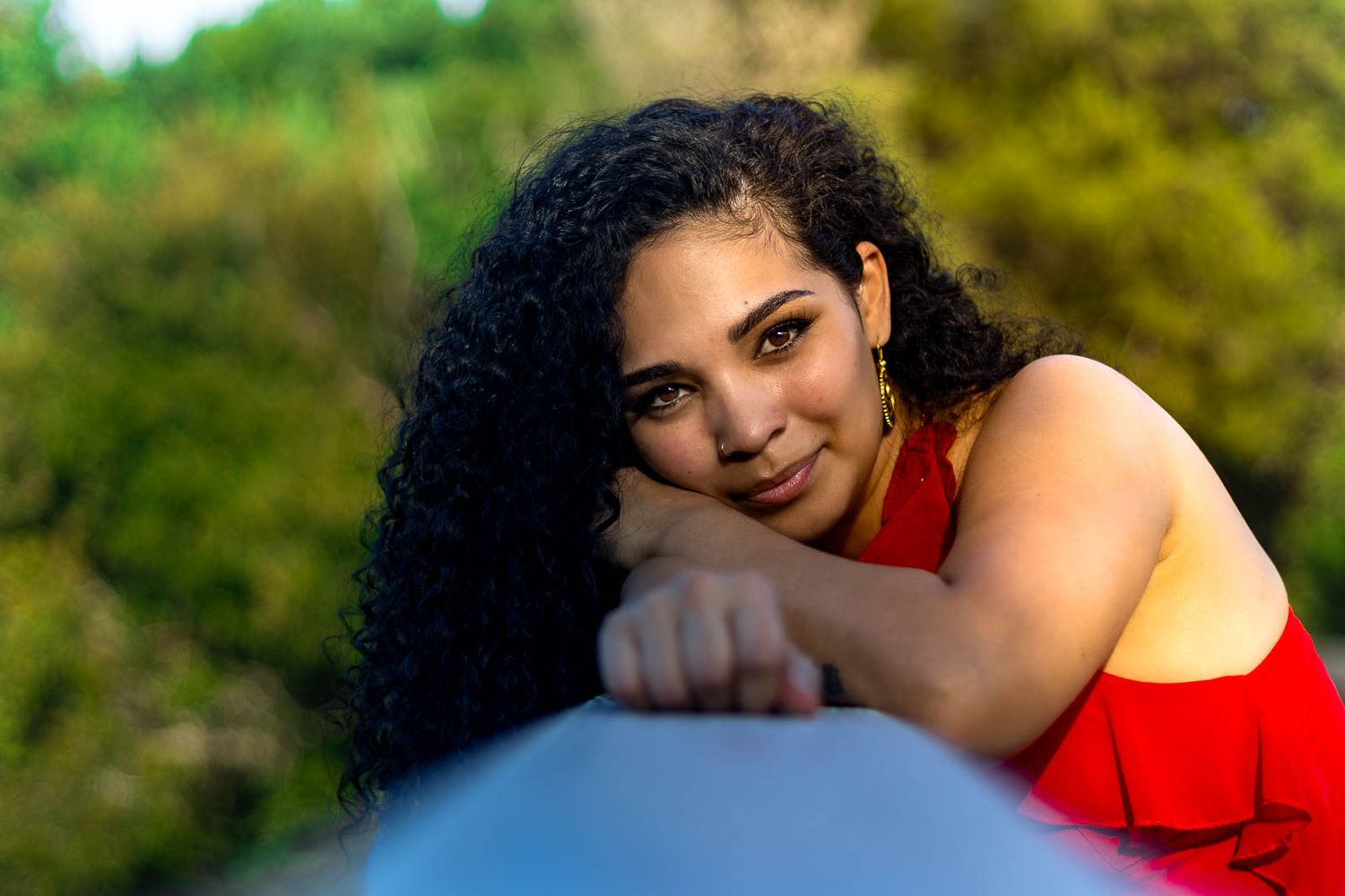 Young woman with curly hair in a red dress leaning on a railing outdoors, smiling warmly with greenery behind her.