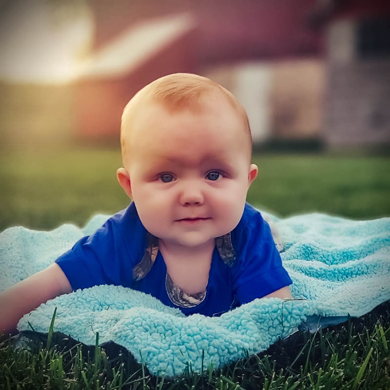 A cute baby in a blue shirt lying on a teal blanket in green grass, looking at the camera.