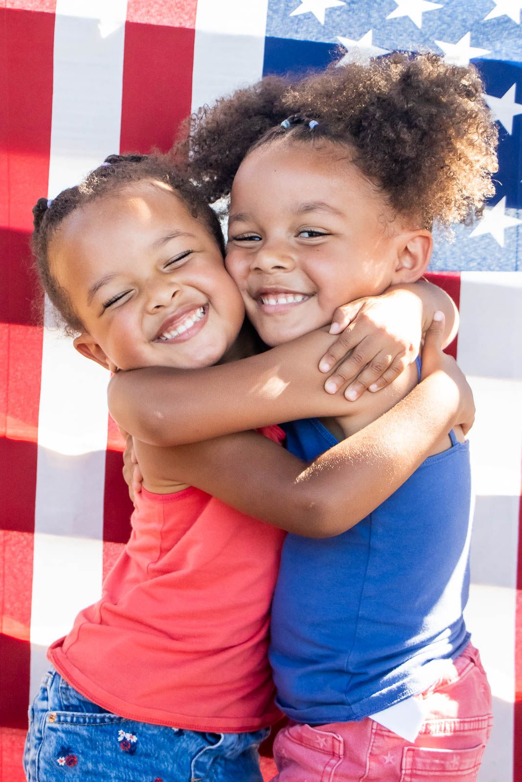 Two young girls hugging and smiling in front of an American flag backdrop.