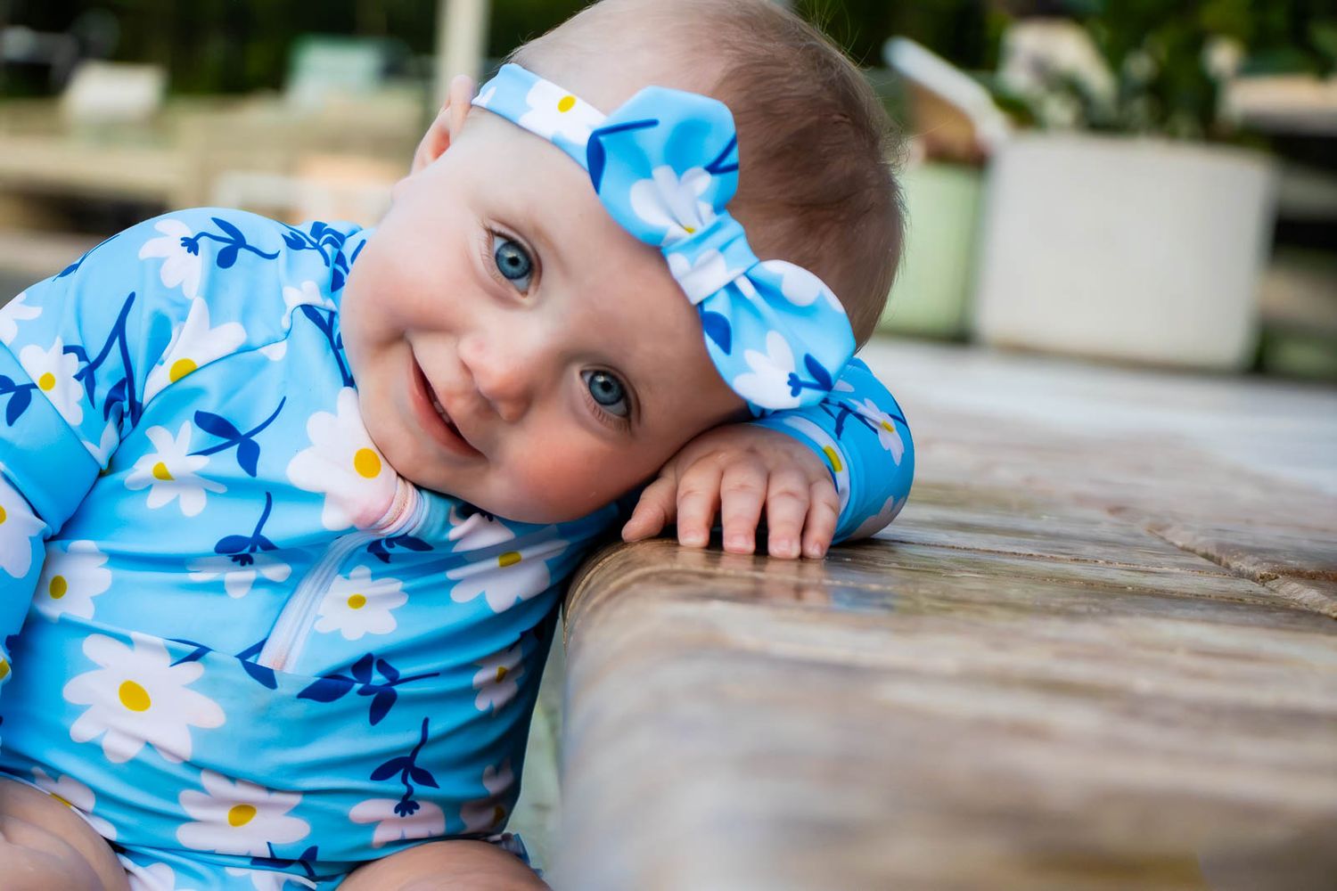 Smiling baby girl in blue daisy floral swimsuit and matching bow headband resting on wooden surface outdoors.