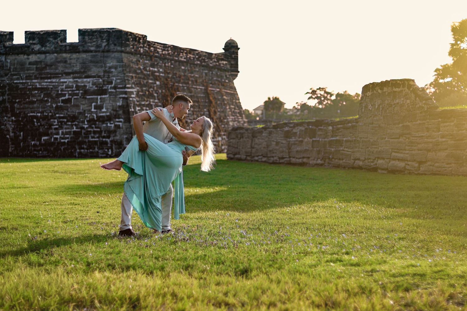 Couple dancing romantically at sunset near historic stone fort ruins on green grass in golden hour light.