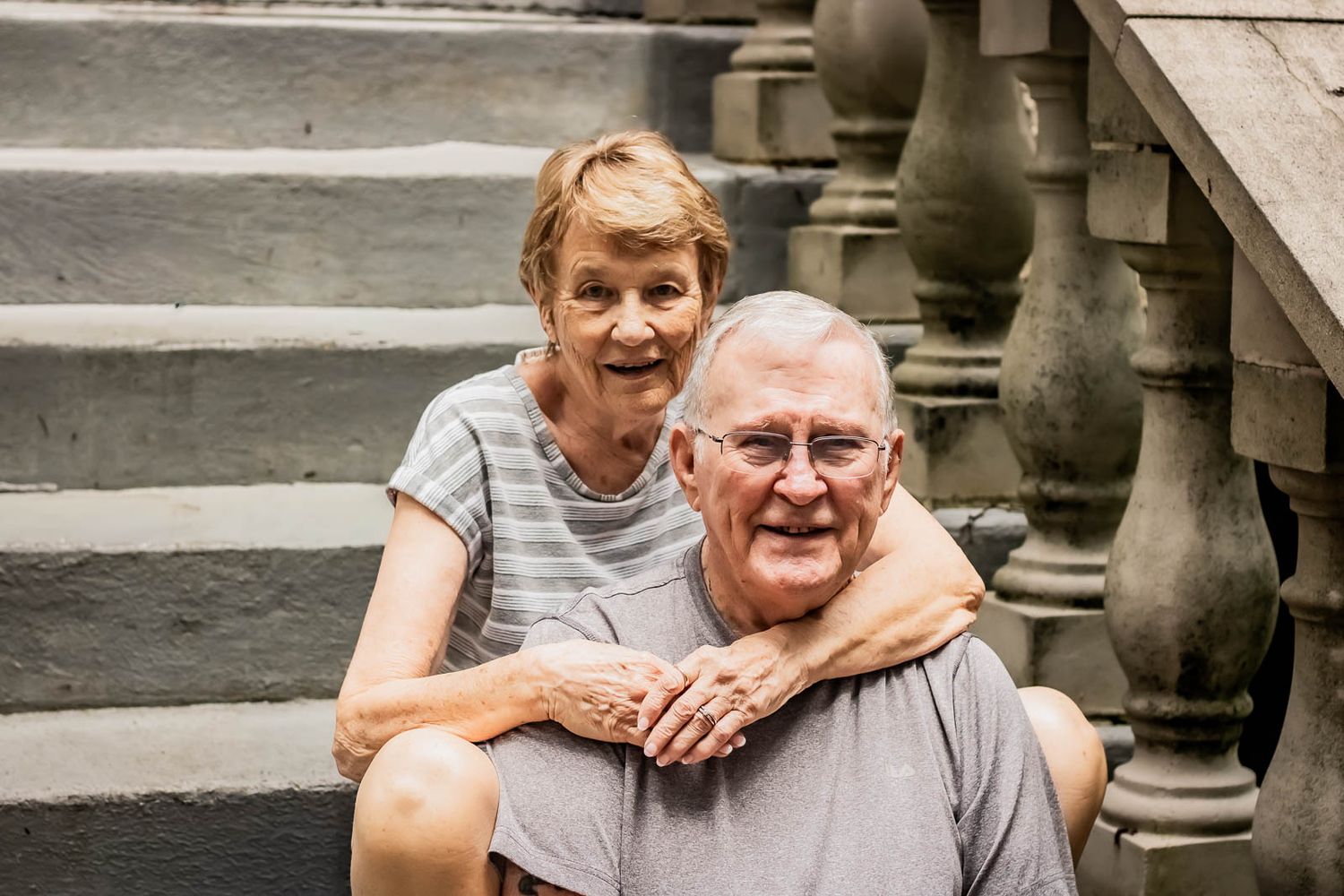 Elderly couple smiling on stone steps, woman embracing man from behind in a warm outdoor portrait.