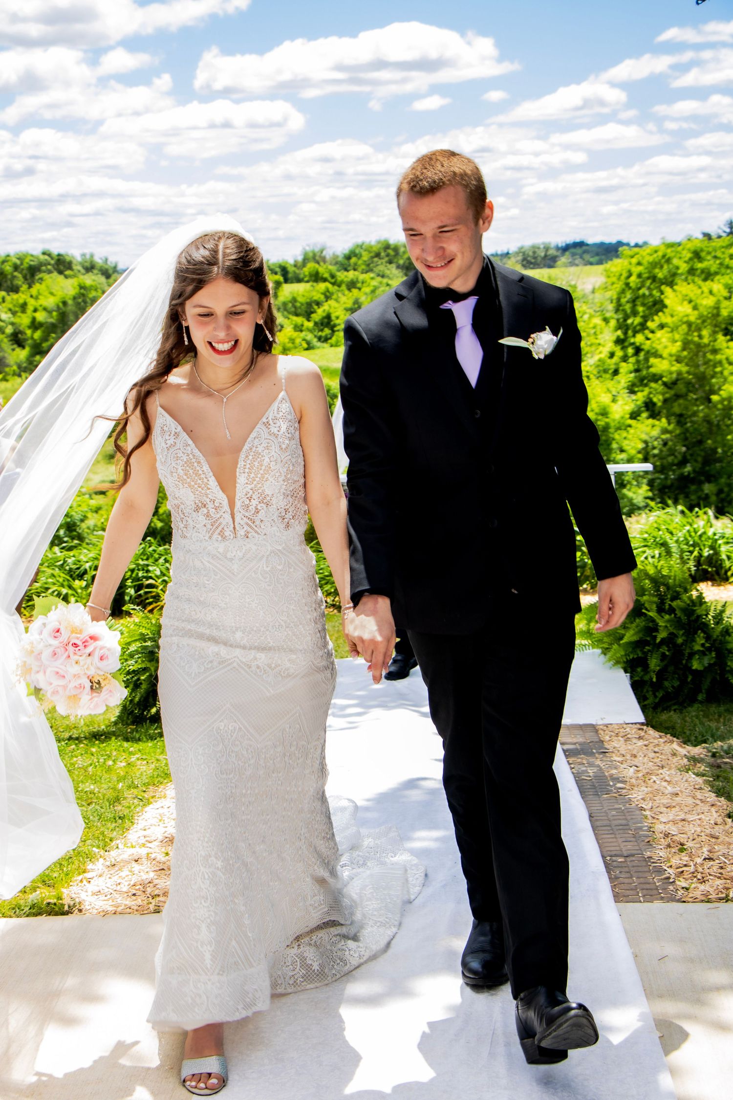 Bride and groom walking down aisle outdoors on sunny day, bride in lace gown holding pink bouquet.