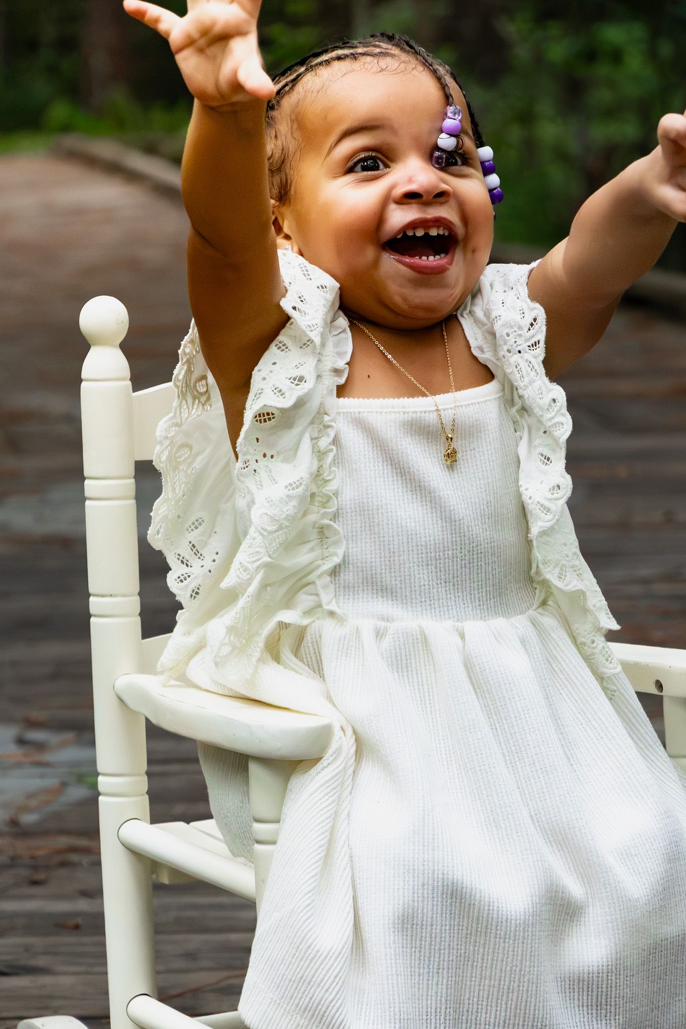 Joyful toddler girl in white dress with purple hair clips raises arms while sitting in white rocking chair outdoors.