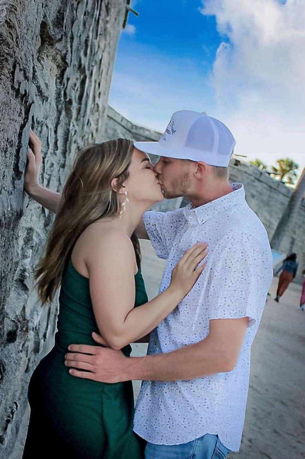 Couple sharing a romantic kiss against a stone wall outdoors, woman in green dress, man in white cap and shirt.