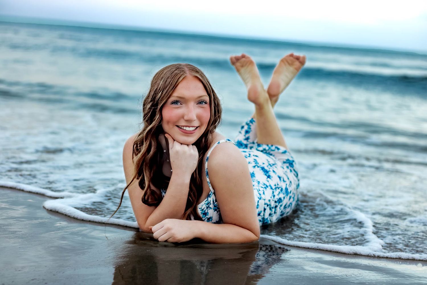 Young woman in blue floral swimsuit posing on sandy beach shore with ocean waves, smiling at camera.