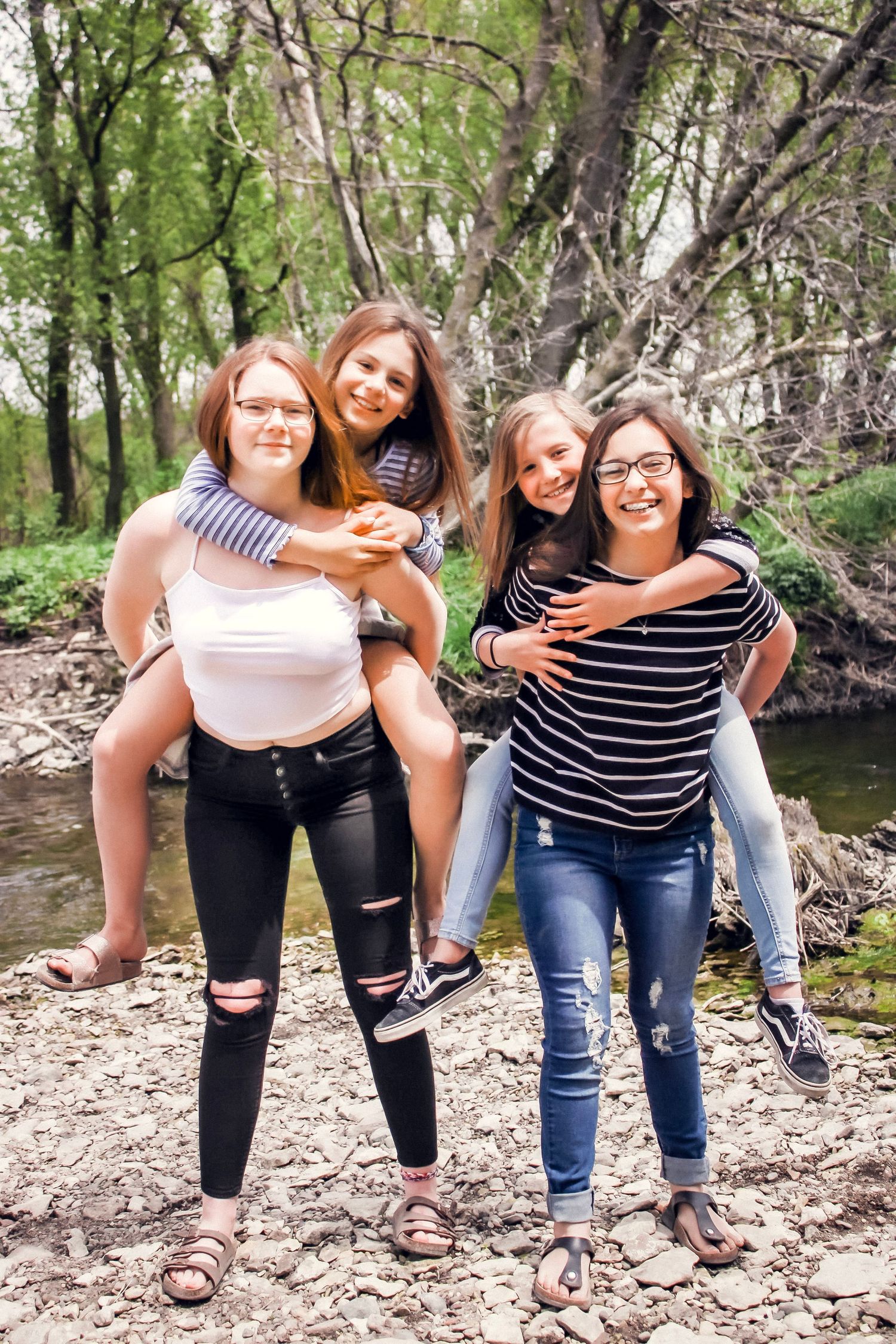 Four sibling girls two standing while the other two are on their backs smiling next to a creek.