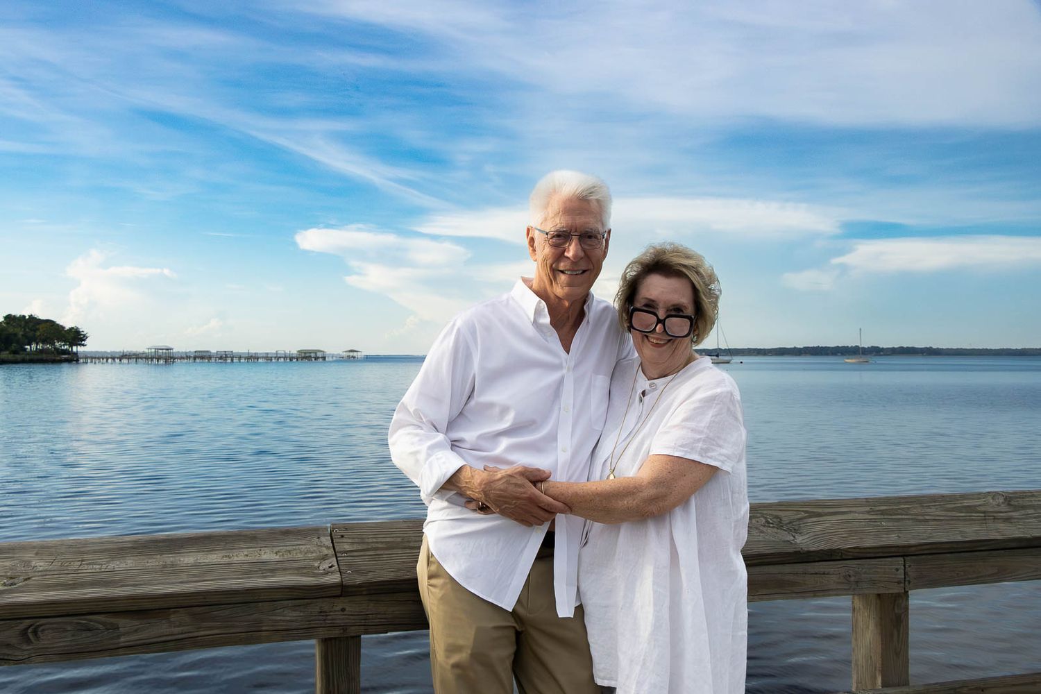 Elderly couple in white shirts embracing on a wooden dock with blue water and sky in the background during their family photography session.