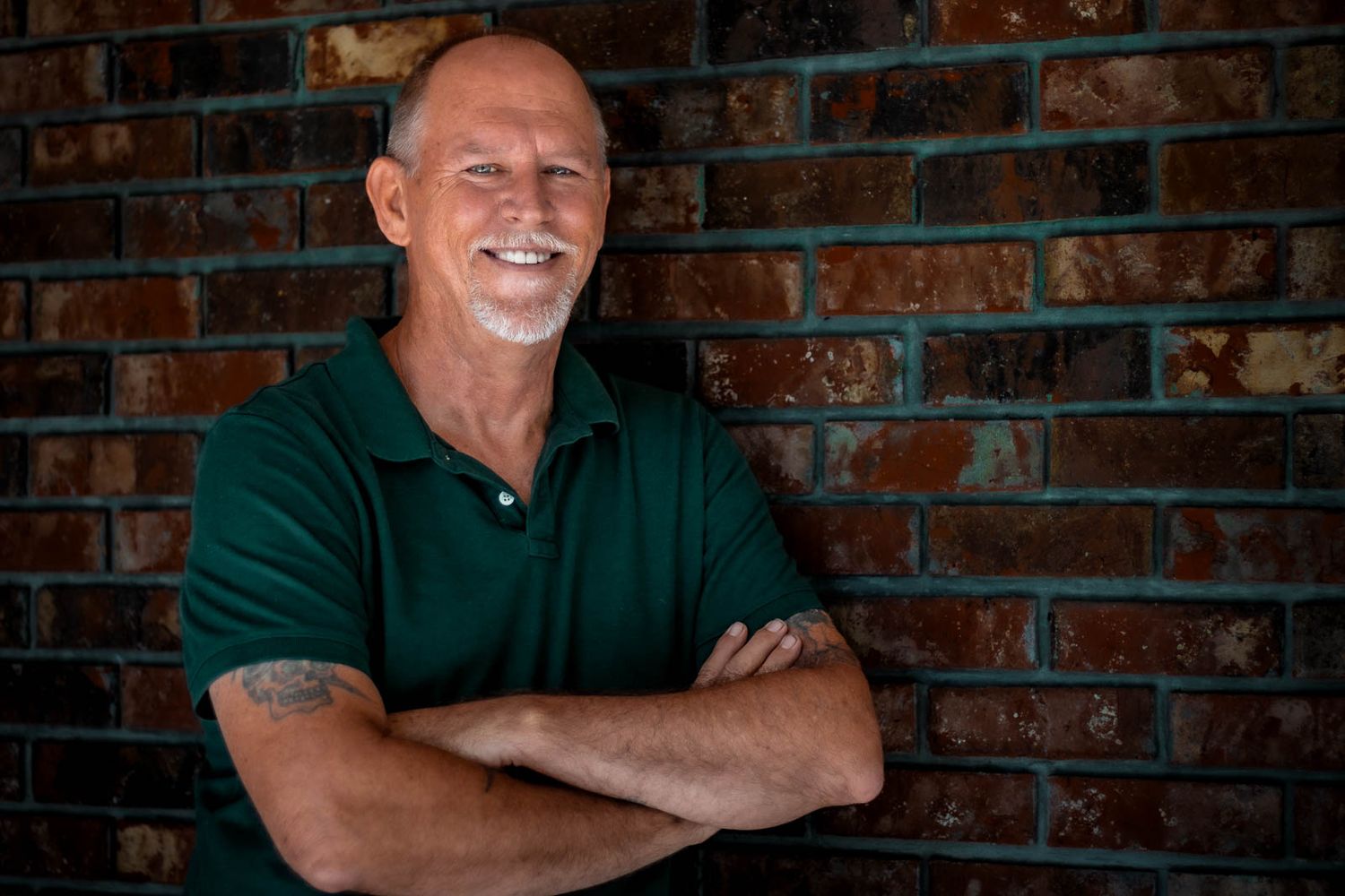 Smiling middle-aged man with arms crossed, wearing green polo shirt, standing against rustic brick wall for his portrait photo session.