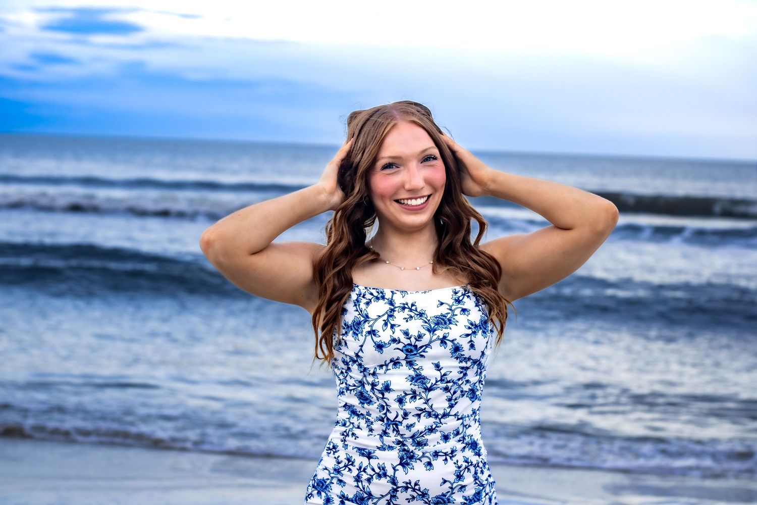 A girl running her hands through her hair standing on the beach with the ocean in the background.