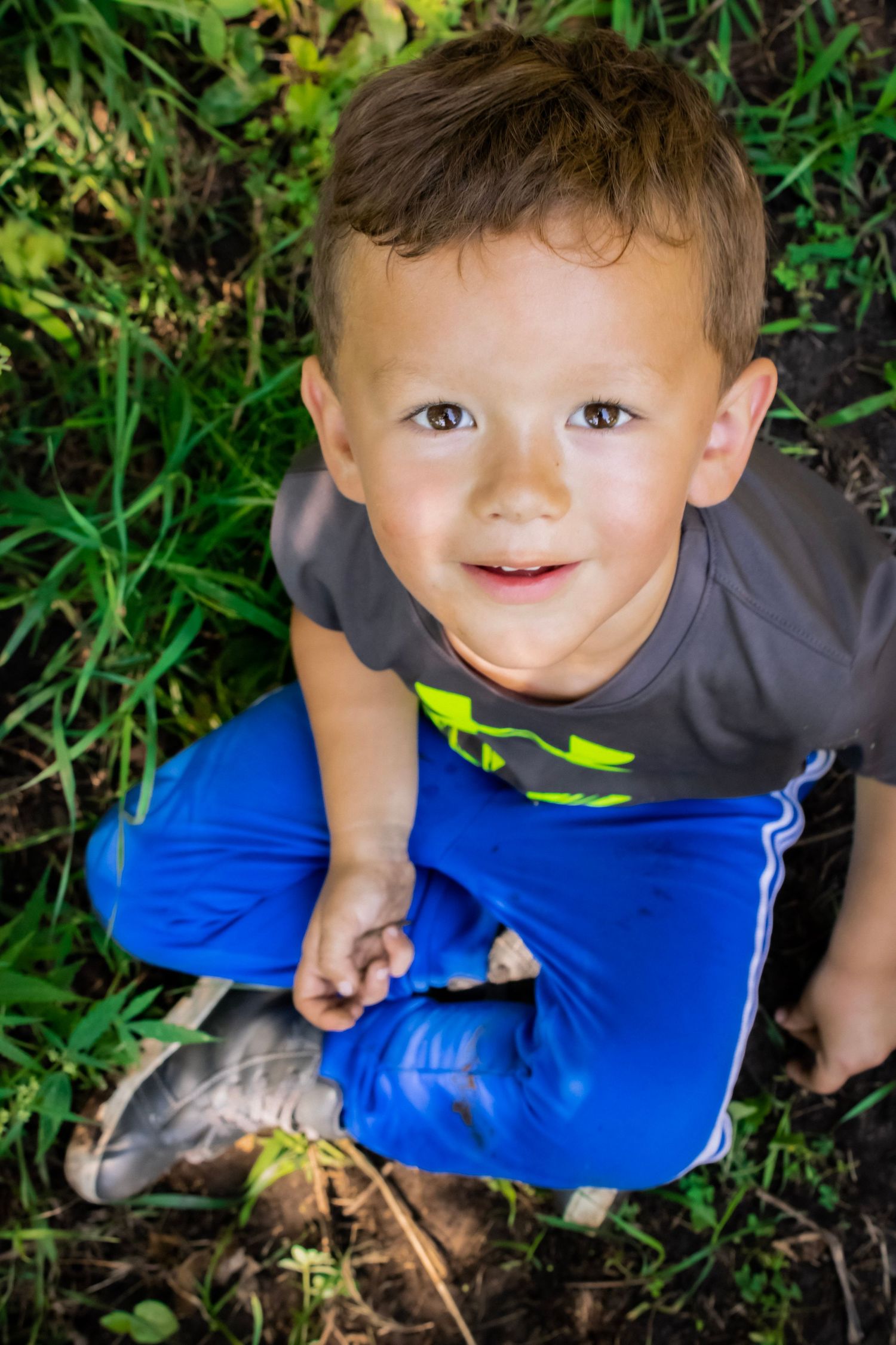 A toddler sitting in the grass and looking up to the sky where the camera is also. This is a birds eye image of the boy.