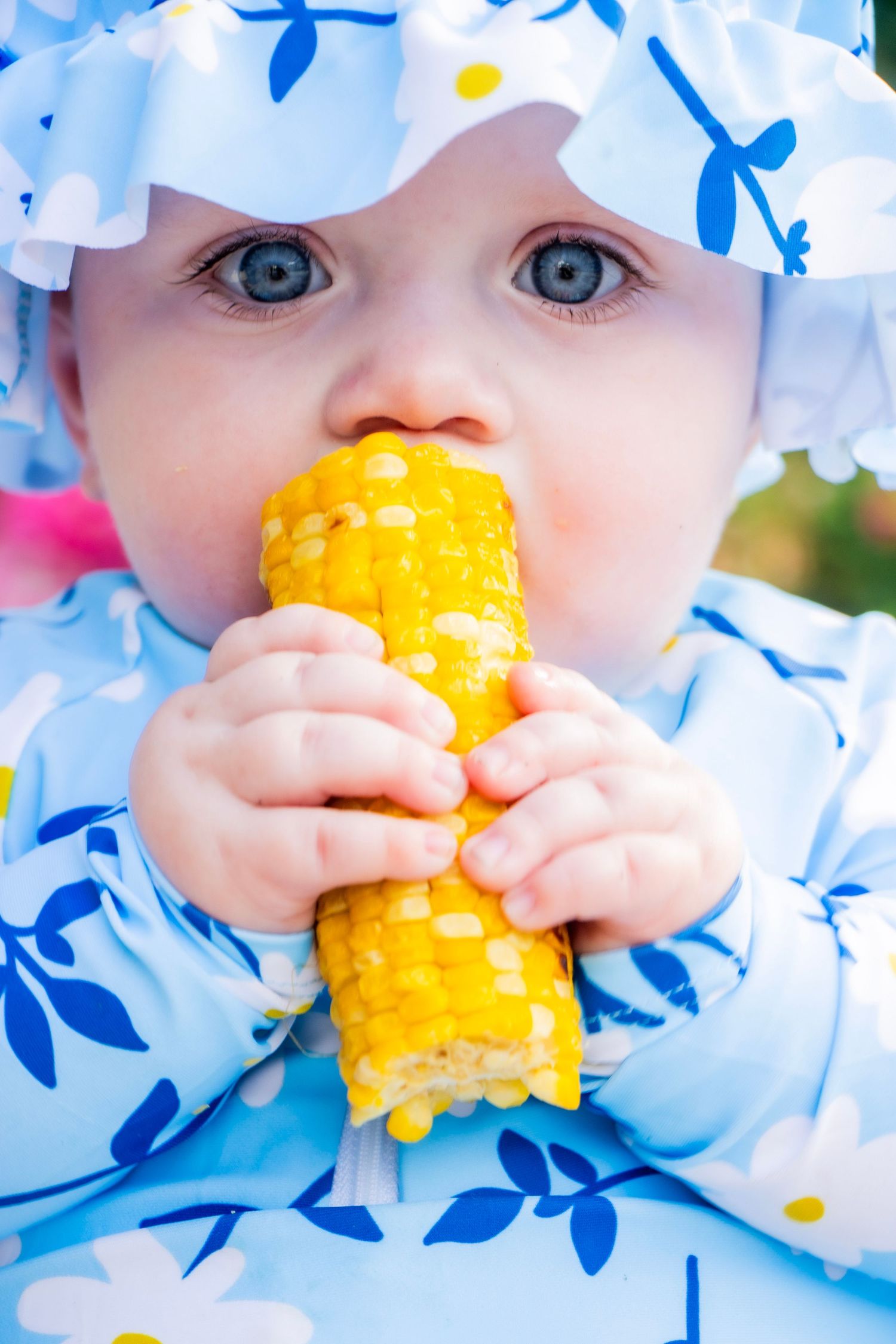A baby girl eating a piece of corn with vibrant blue and yellow colors smiling.