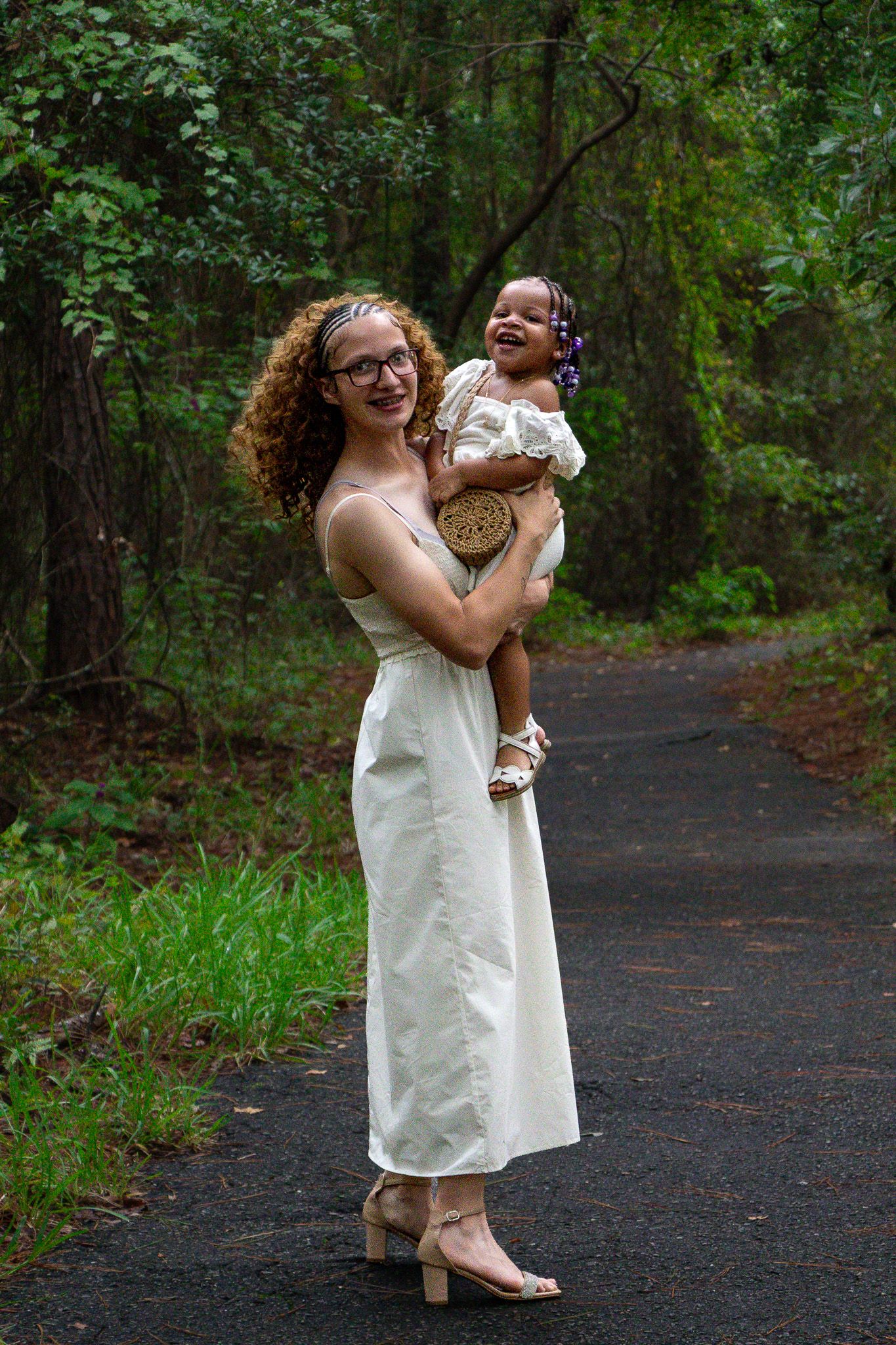 A smiling woman in a white dress holds a laughing toddler on a forest path surrounded by lush green trees.