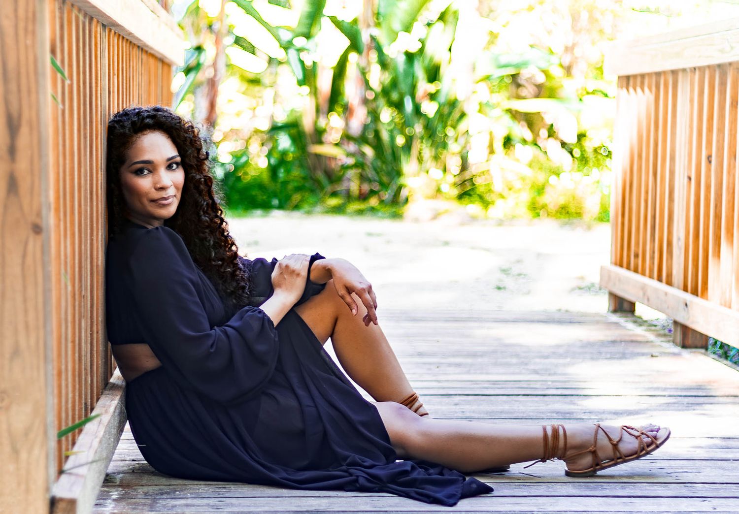 Young woman in sitting on wooden boardwalk surrounded by tropical greenery in outdoor portrait session in crescent beach, fl.