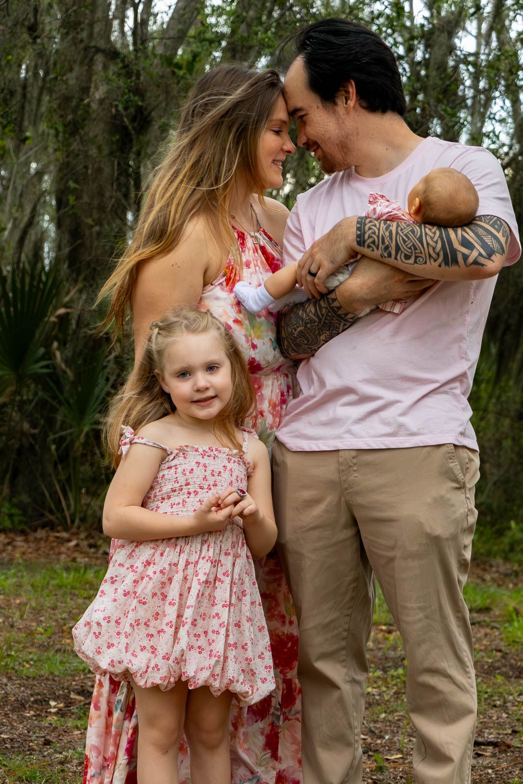 Family portrait of parents with tattooed dad holding newborn and young daughter in floral dress outdoors.