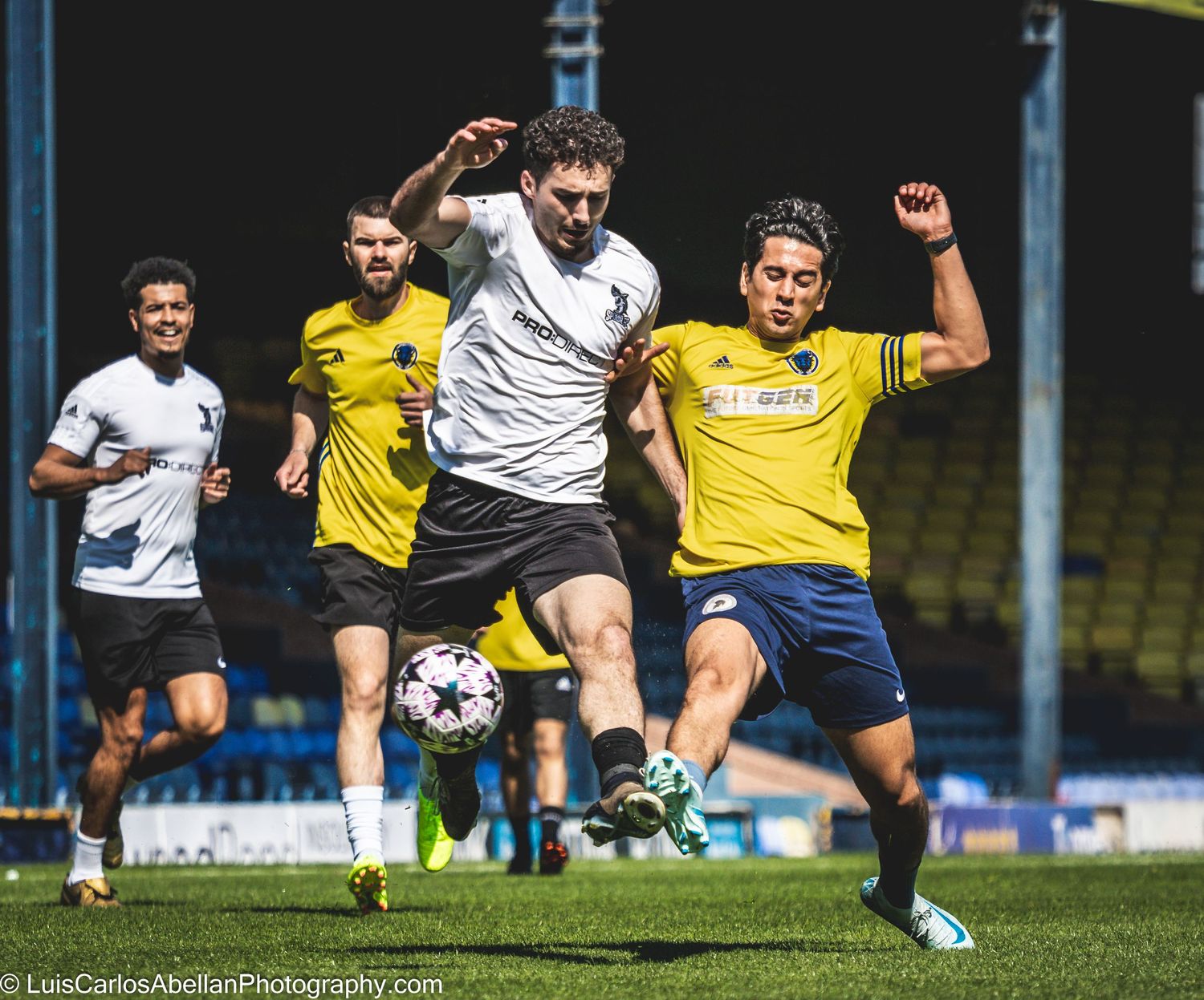 Dynamic matchday action in Southeast England: a white kit player kicks the ball while challenging an opponent, by Luis Carlos Abellan.