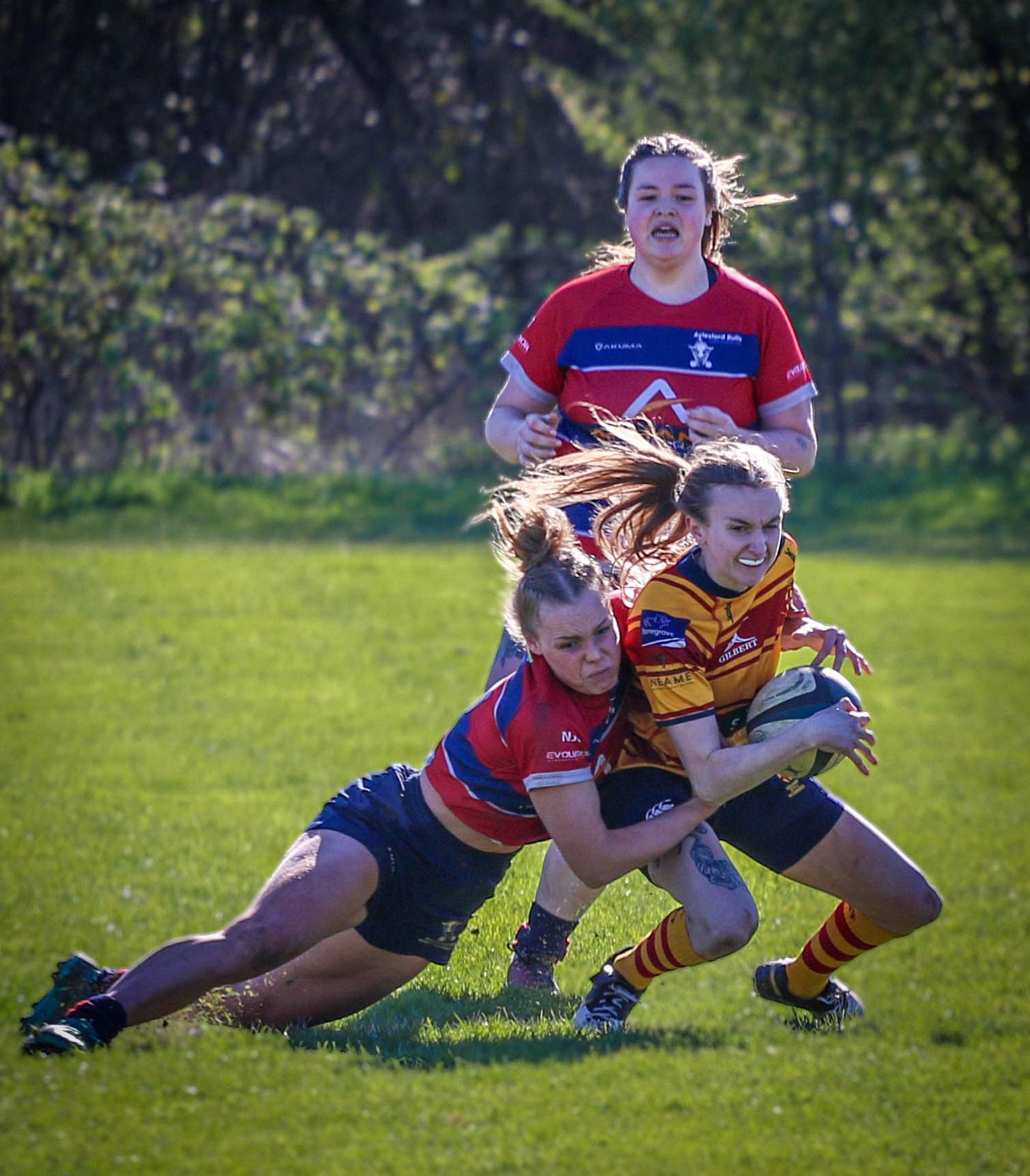 A female rugby player in red and blue tackles an opponent in gold stripes during a match in Aylesford, by Luis Carlos Abellan Photography