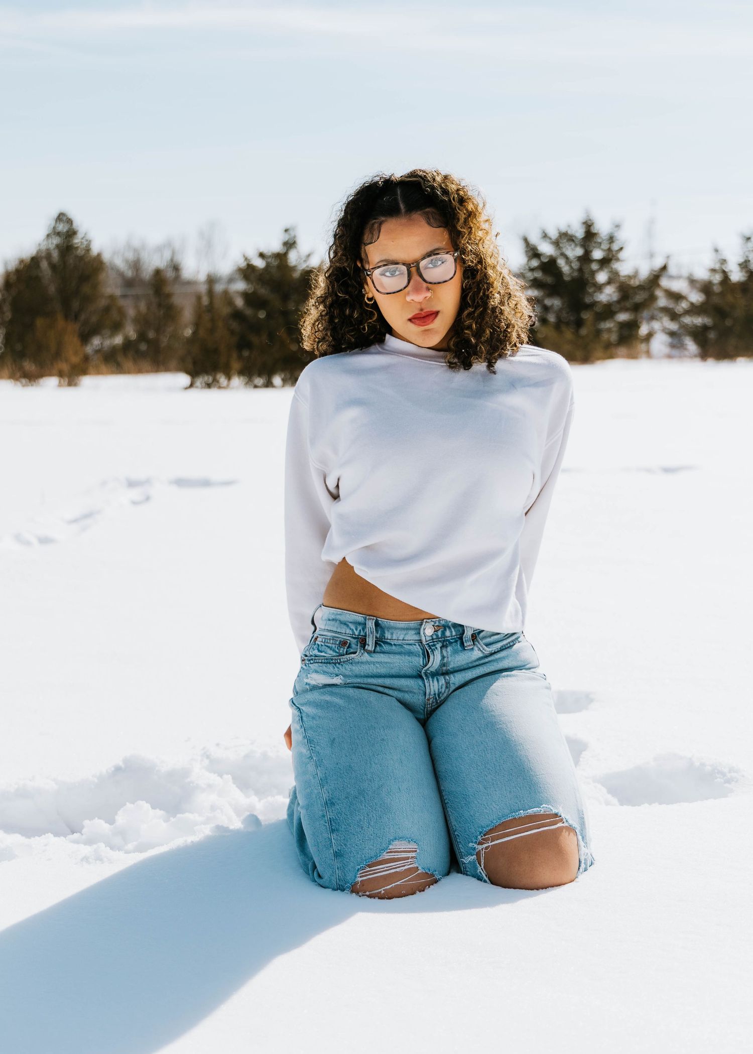 High school senior in a white sweater and jeans with glasses is a snow covered field in St. Robert, MO for her senior phot session
