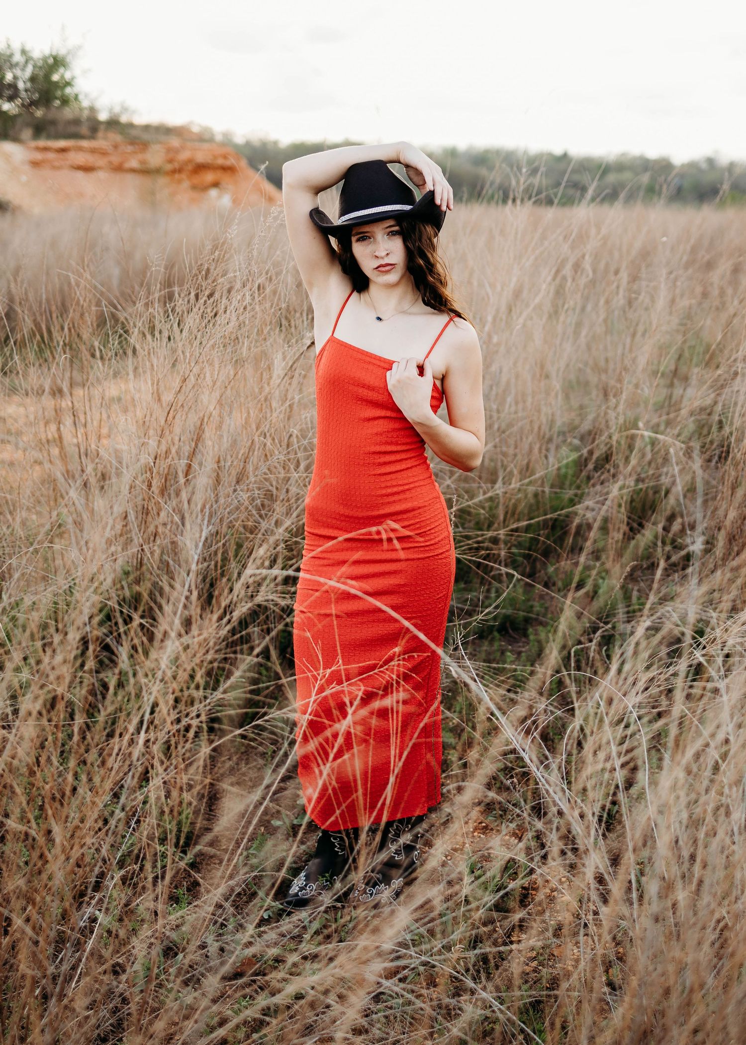 high school senior in a cowboy hat, boots and a red dress in a field for her senior session in Waynesville, MO.
