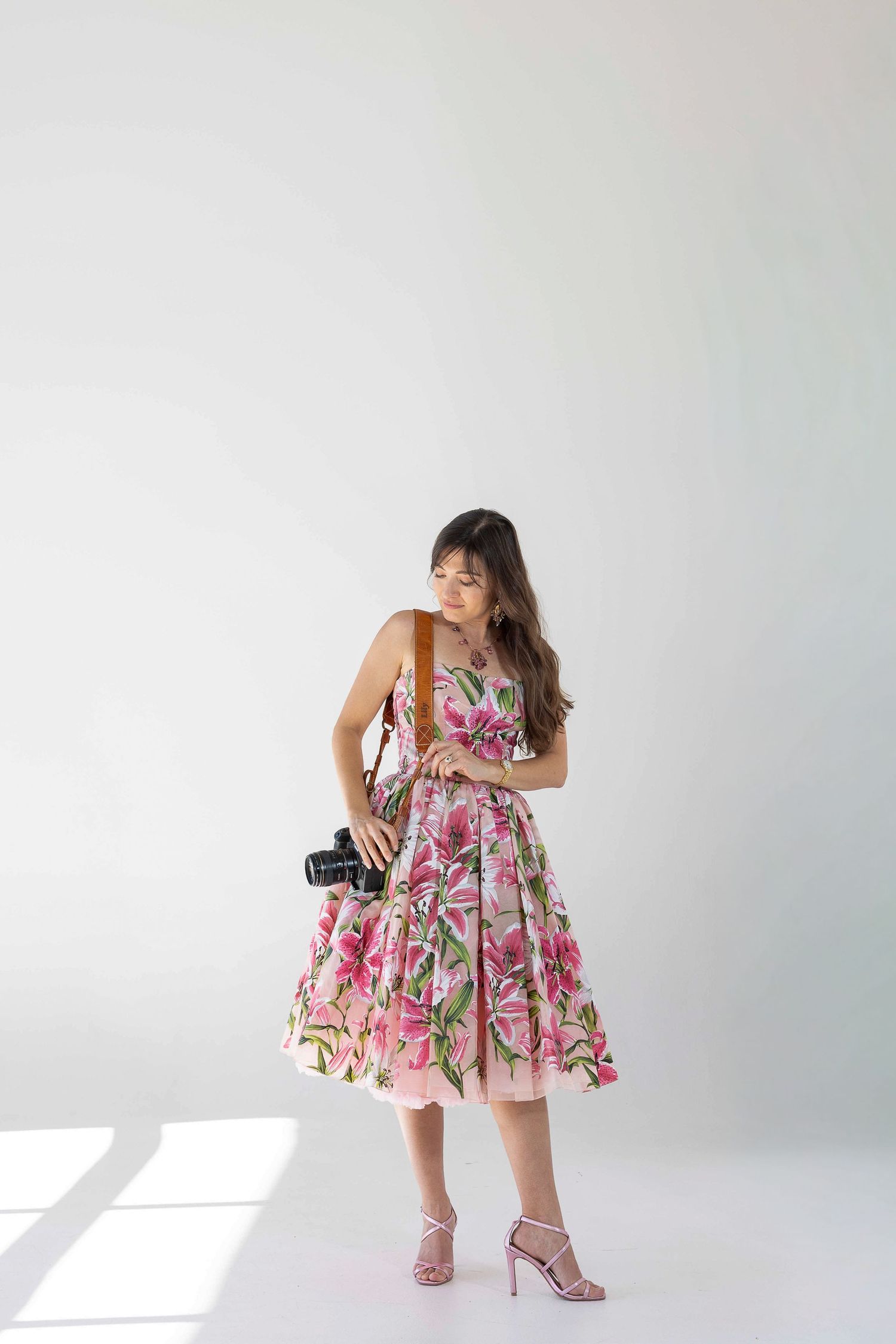 Woman in floral midi dress holding camera, wearing pink heeled sandals, posing in bright studio setting.