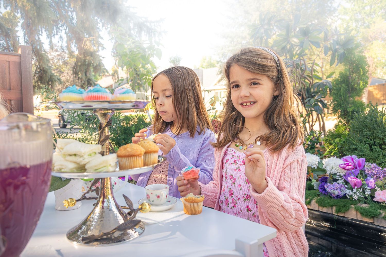 Two young girls enjoy a garden tea party with cupcakes on a tiered stand, teacups, and a pitcher of pink drink surrounded by purple flowers.