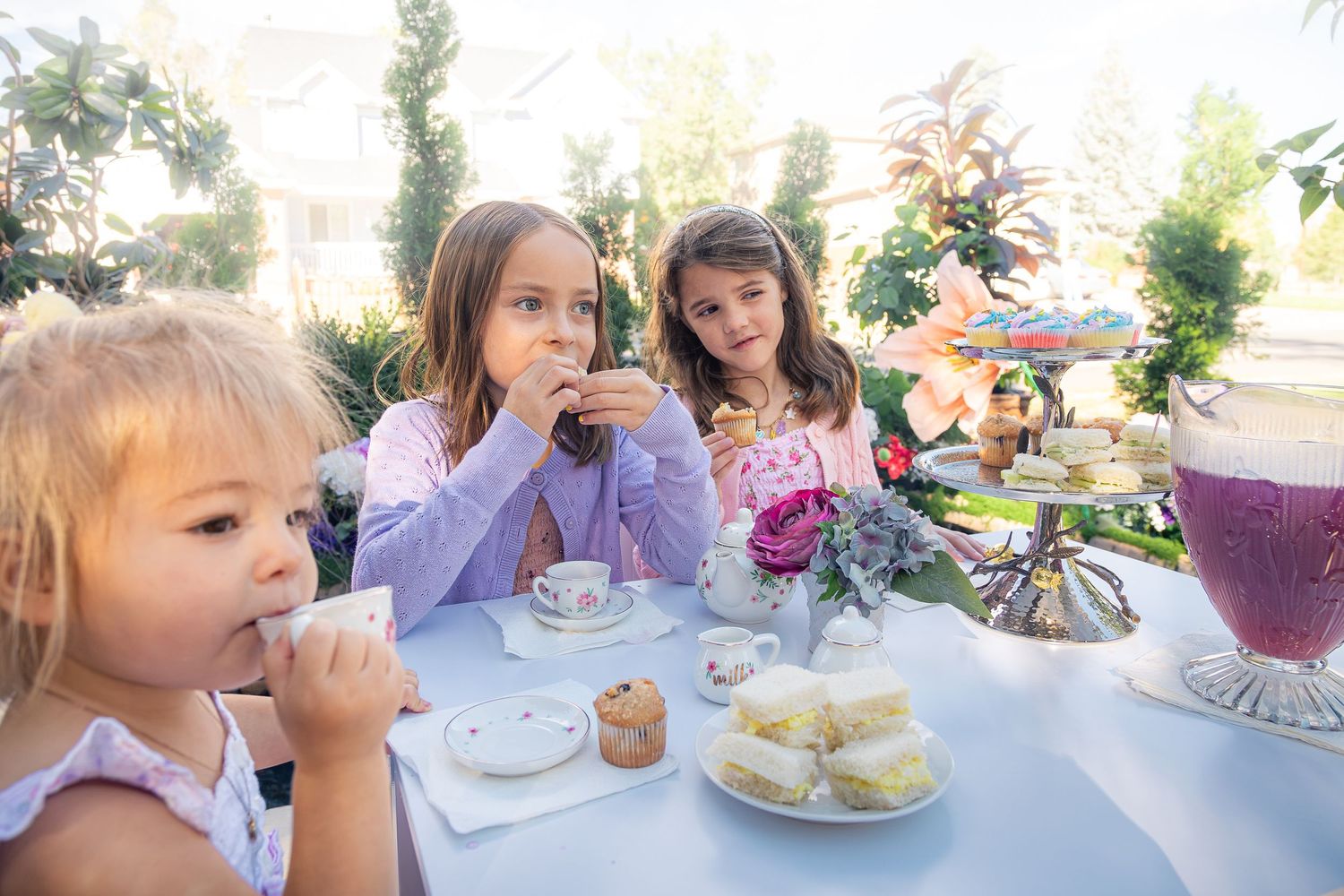 Three young girls enjoy a garden tea party with teacups, sandwiches, a pink drink and a tiered dessert stand surrounded by flowers.