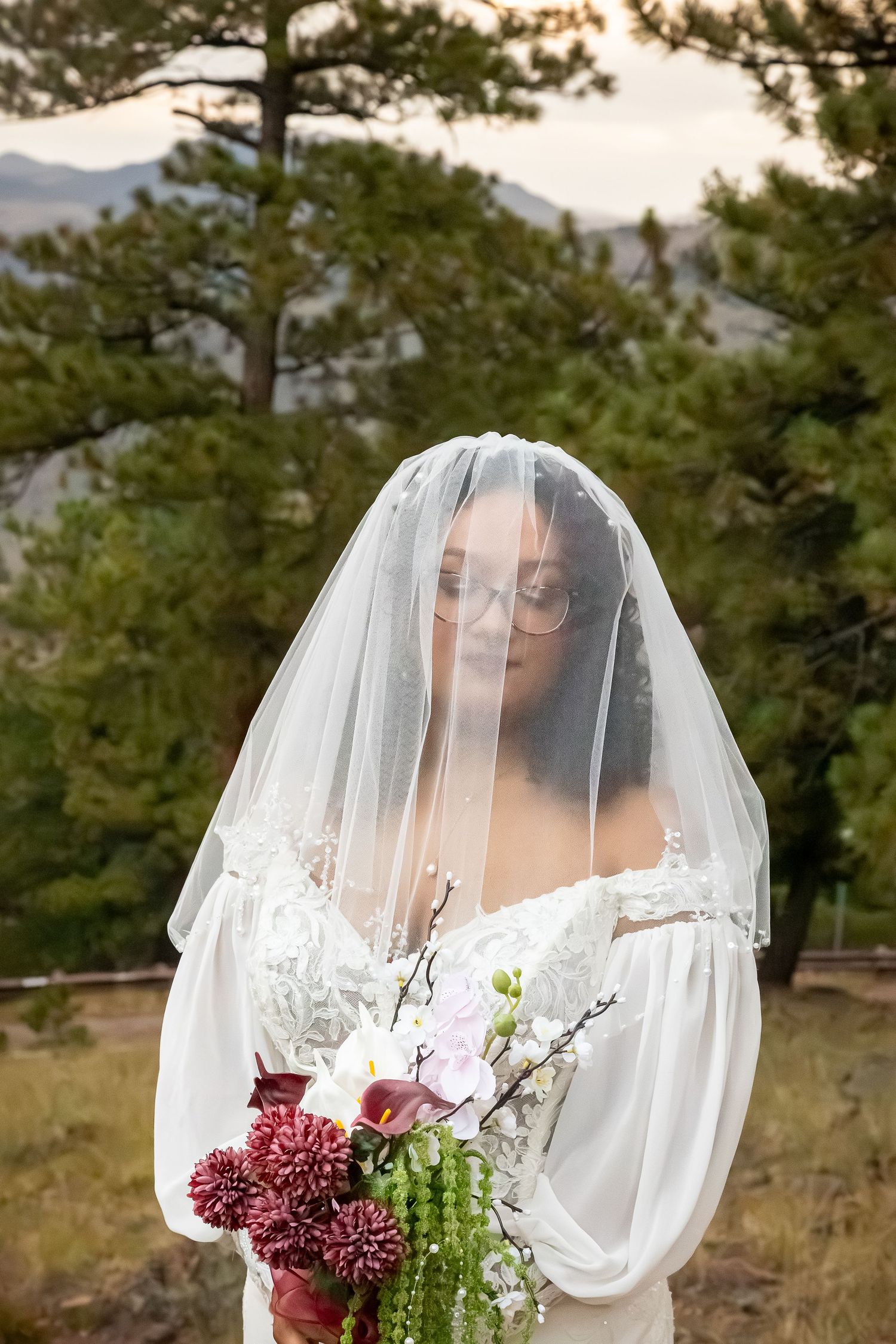 Bride in white gown and veil holds floral bouquet outdoors with pine trees and mountains in background.