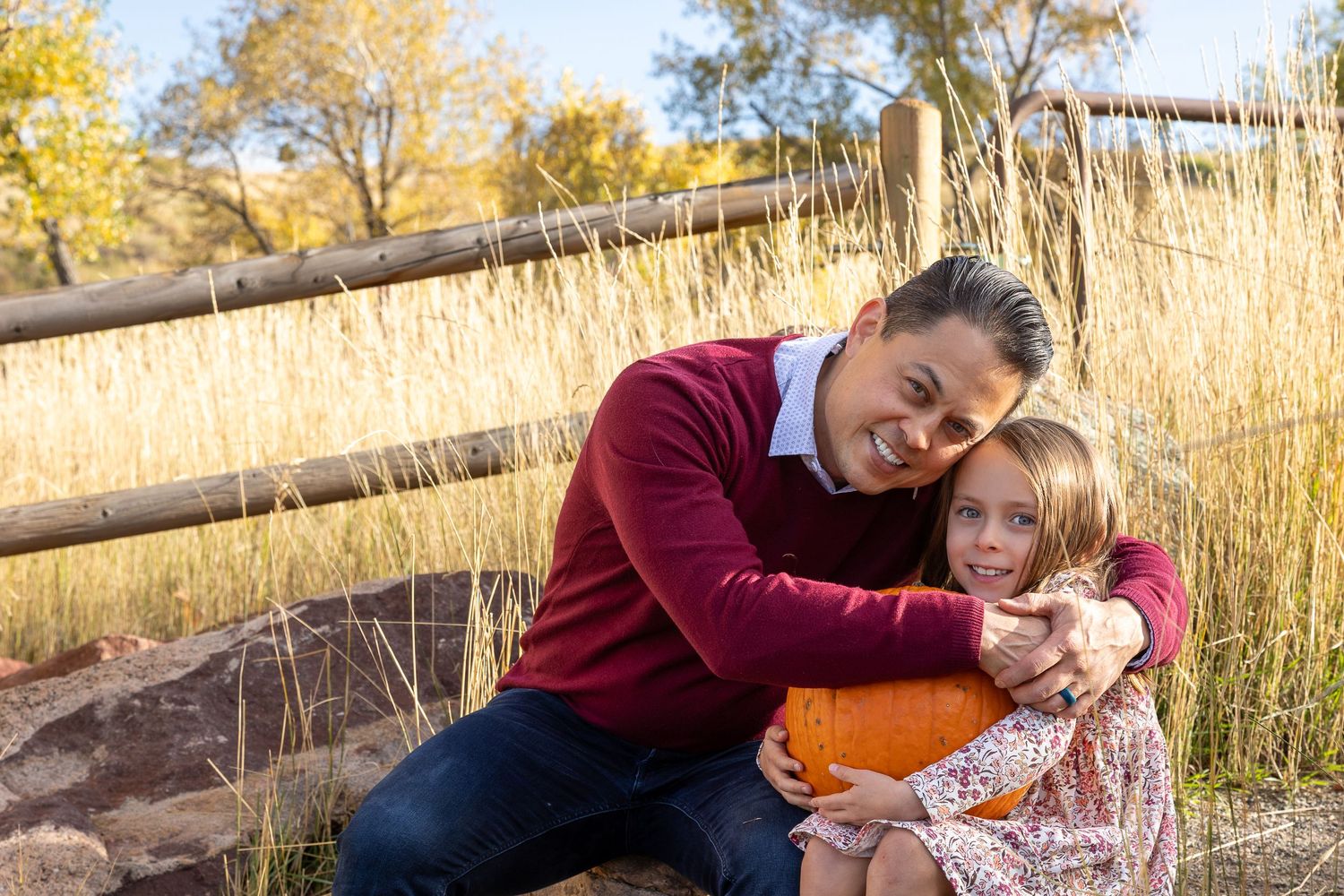 A father and daughter share a warm hug outdoors in autumn, holding a pumpkin near a rustic wooden fence.