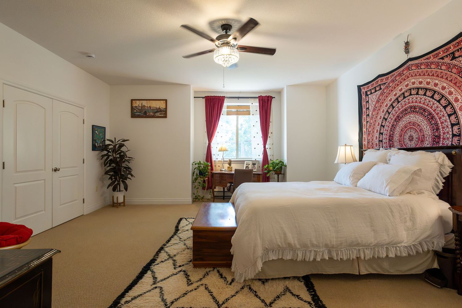 Spacious bedroom with white bedding, ceiling fan, red mandala tapestry, patterned rug, and bright window with red curtains.
