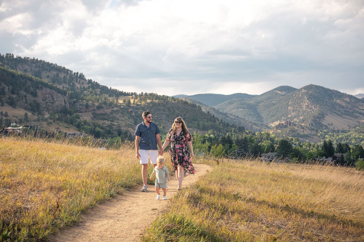 Family of three walking on a mountain trail through golden meadows with scenic Colorado foothills in the background.