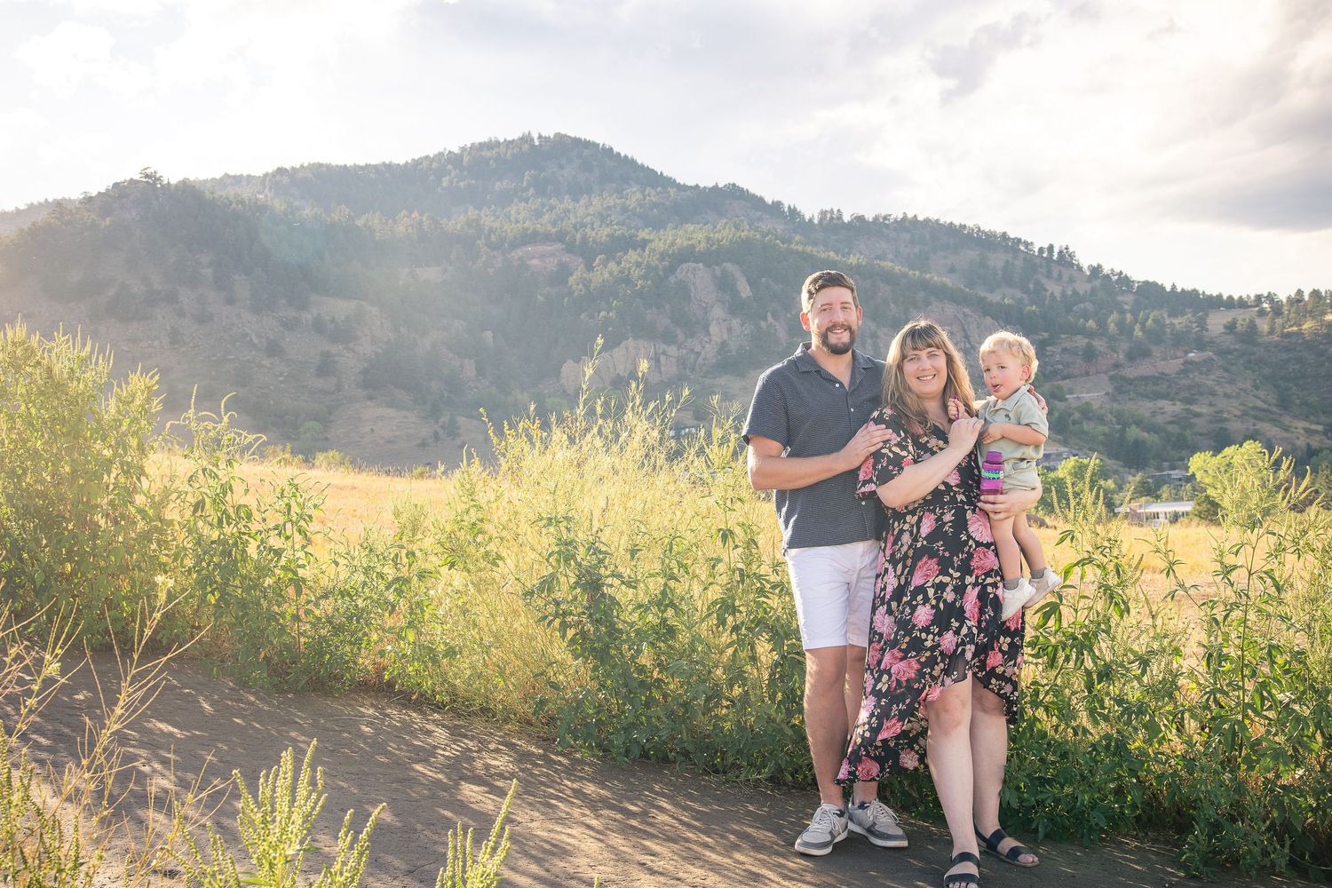 Family of three posing on a dirt path with green meadows and a sunlit mountain backdrop during golden hour.