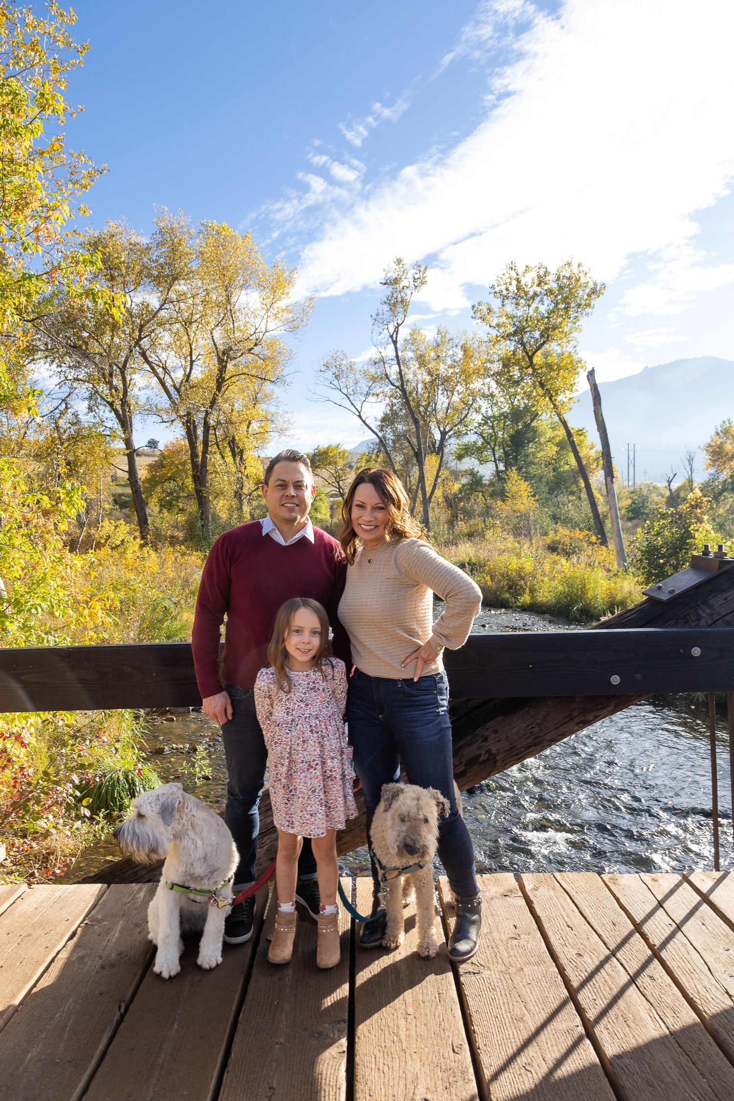 Family of three with two dogs posing on a wooden bridge surrounded by autumn trees and blue sky.