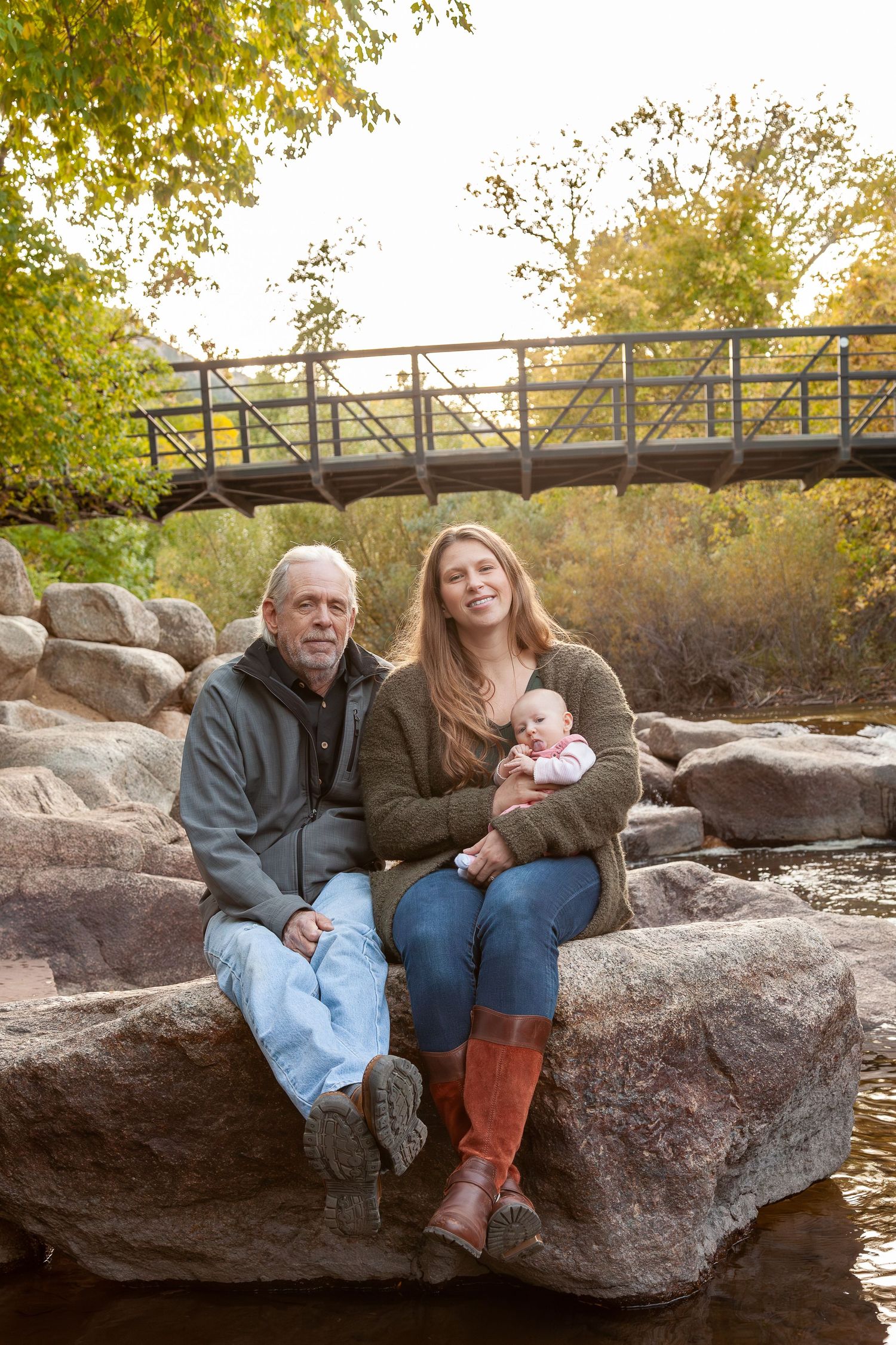 Grandpa and young mother holding newborn baby sit on rocks near a bridge in autumn park.