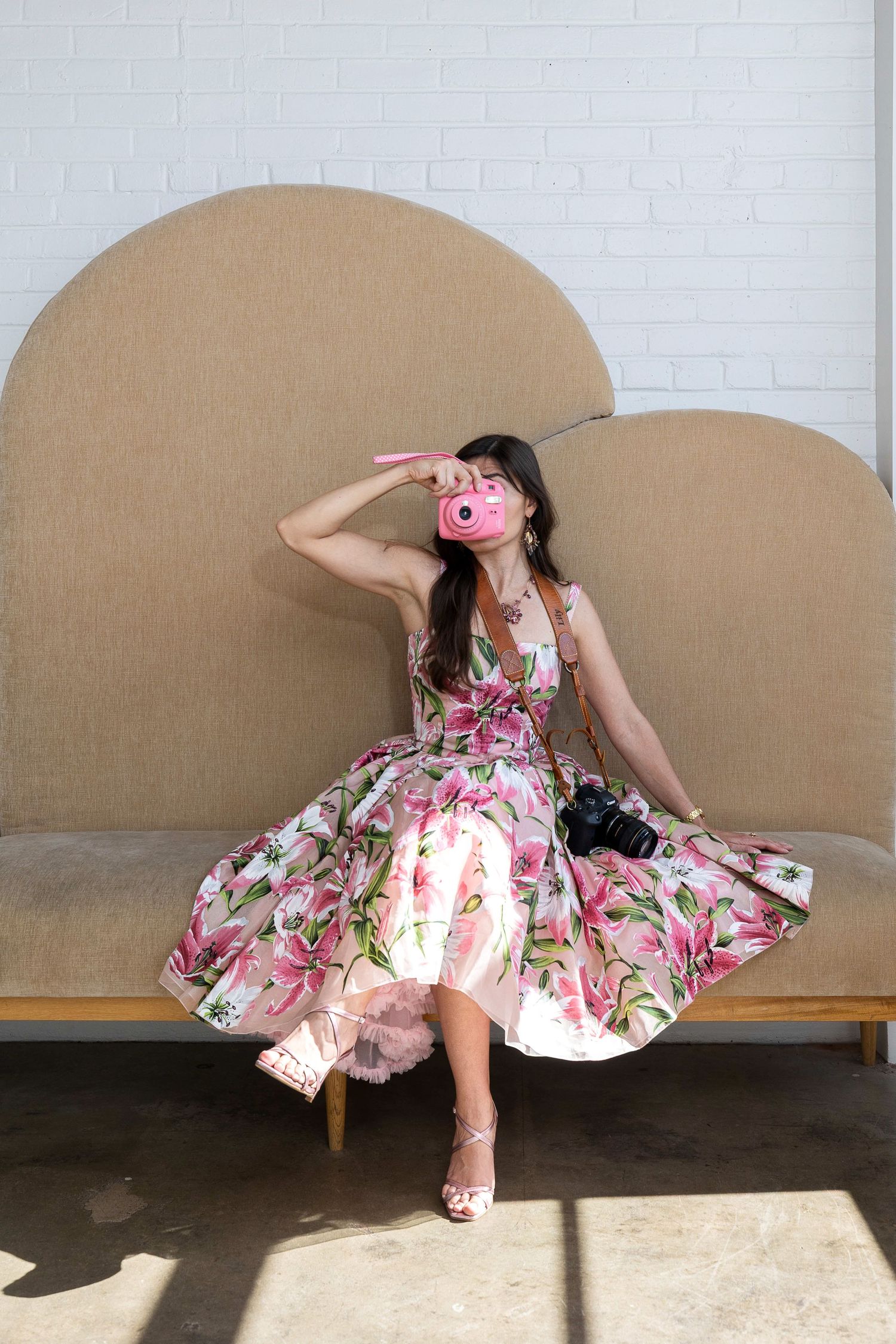 Woman in floral pink dress sitting on beige sofa, holding pink Instax camera up to her eye.