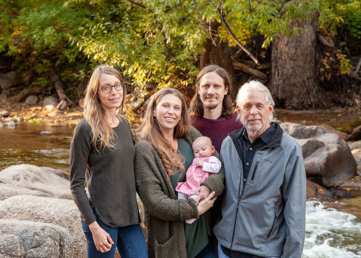 Multi-generational family of five poses outdoors by a rocky river, including a baby, surrounded by autumn trees.