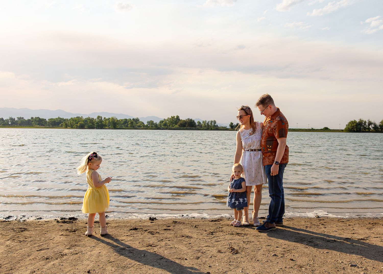 Family of four enjoying a sunny day on a sandy lakeside beach with lush green trees in the background.