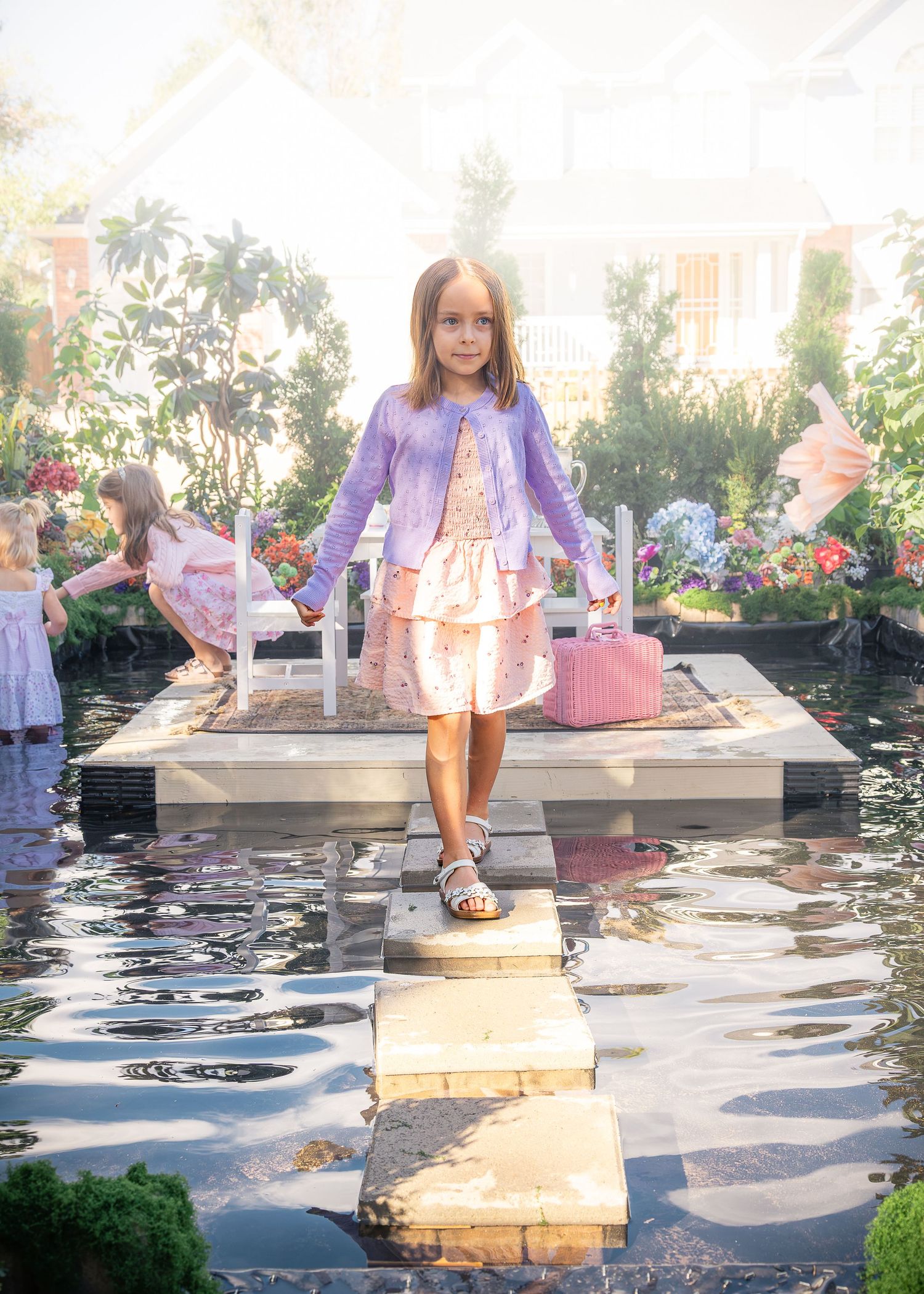 Little girl in a lavender cardigan and pink dress walks across stepping stones over a garden pond with flowers behind her.