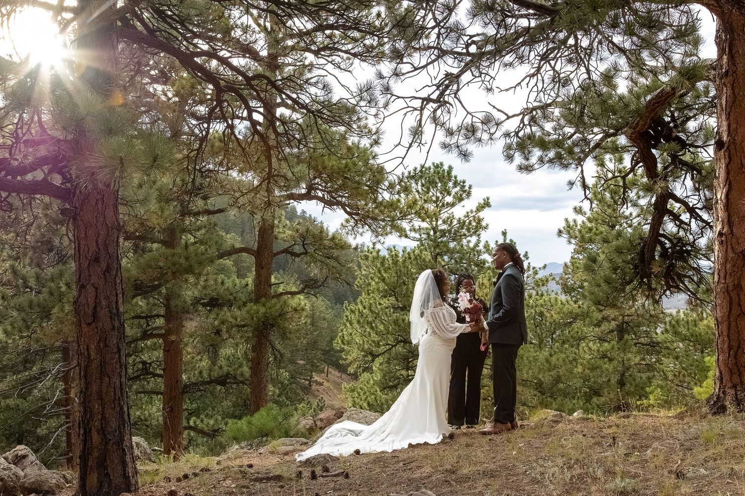 Couple exchanges vows during an outdoor forest wedding ceremony surrounded by pine trees with sunlight streaming through the branches.