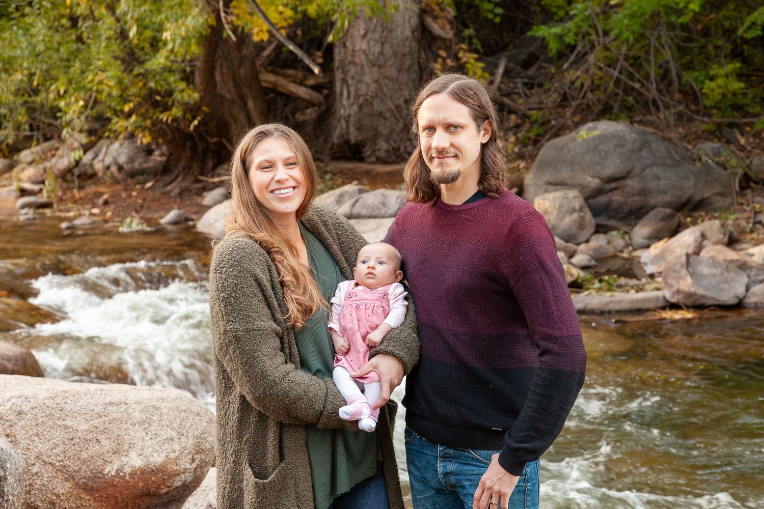 Young family with newborn baby posing by a rocky mountain stream surrounded by autumn foliage.