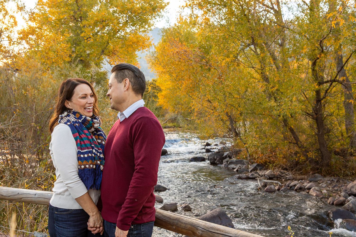 Couple laughing together on a wooden bridge surrounded by vibrant autumn foliage and a flowing river.