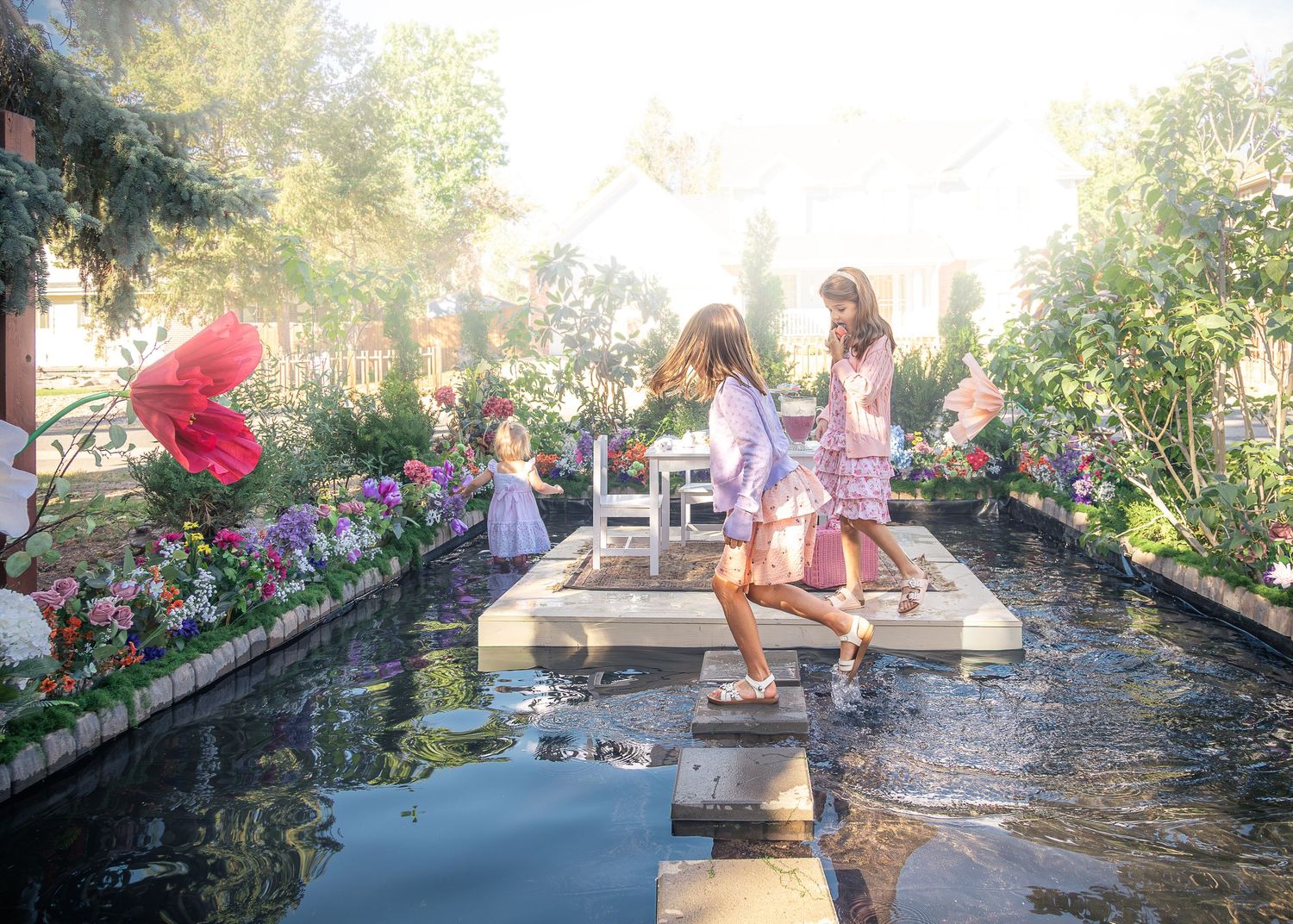 Two girls in pastel dresses walk across stepping stones over a reflective garden pond surrounded by colorful flowers and lush greenery.
