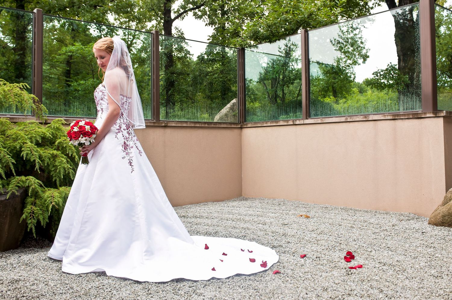 Bride in white gown with veil holds red bouquet, standing in a gravel courtyard with rose petals scattered around.