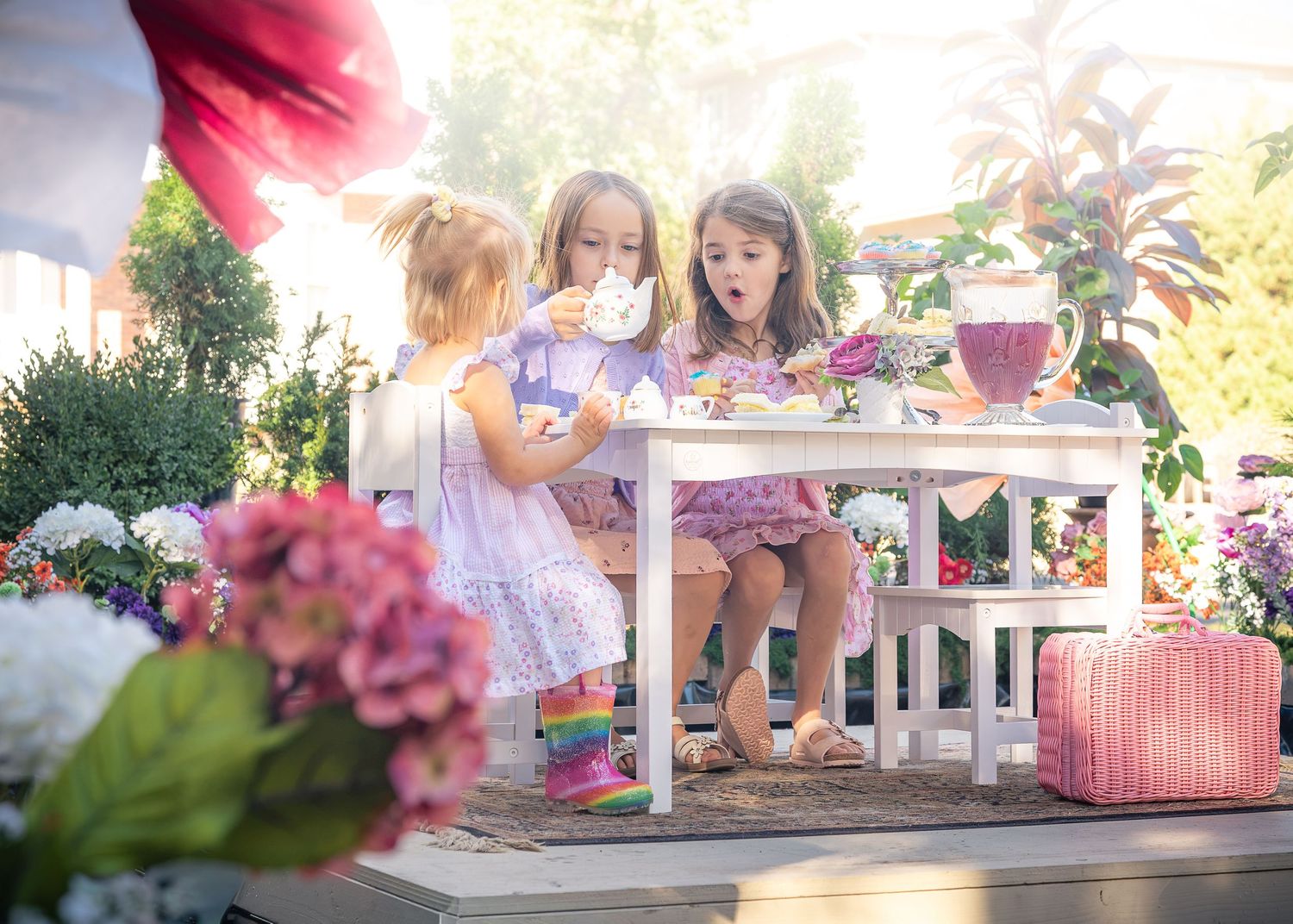 Three little girls in pastel dresses enjoy a tea party at a small white table surrounded by pink and white flowers in a sunny garden.